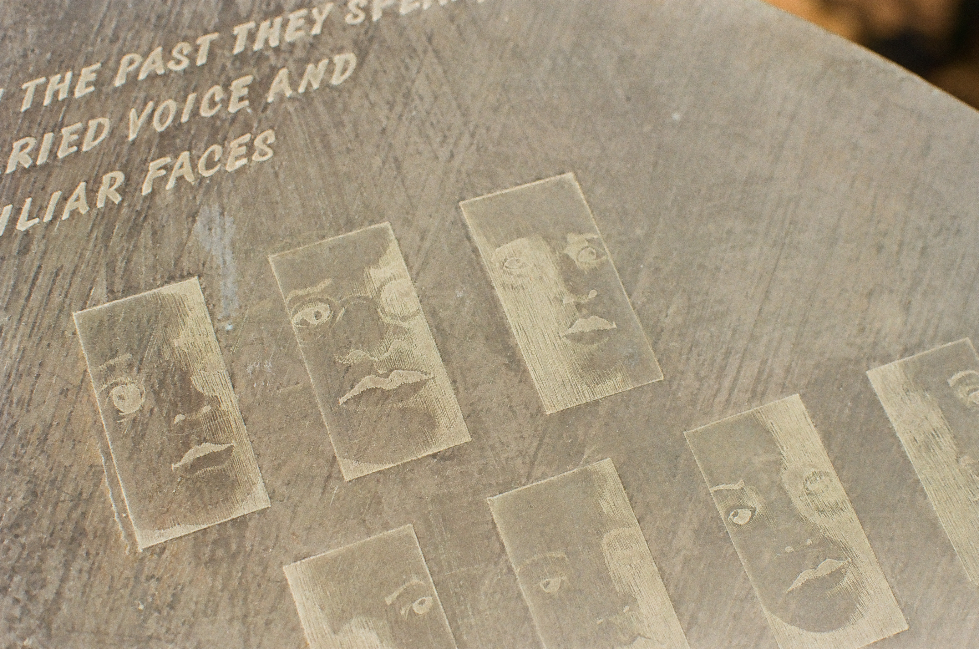 A close view of a memorial stone with carved capital letters reading partial phrases about the past, voice, and familiar faces, above rows of etched face panels.