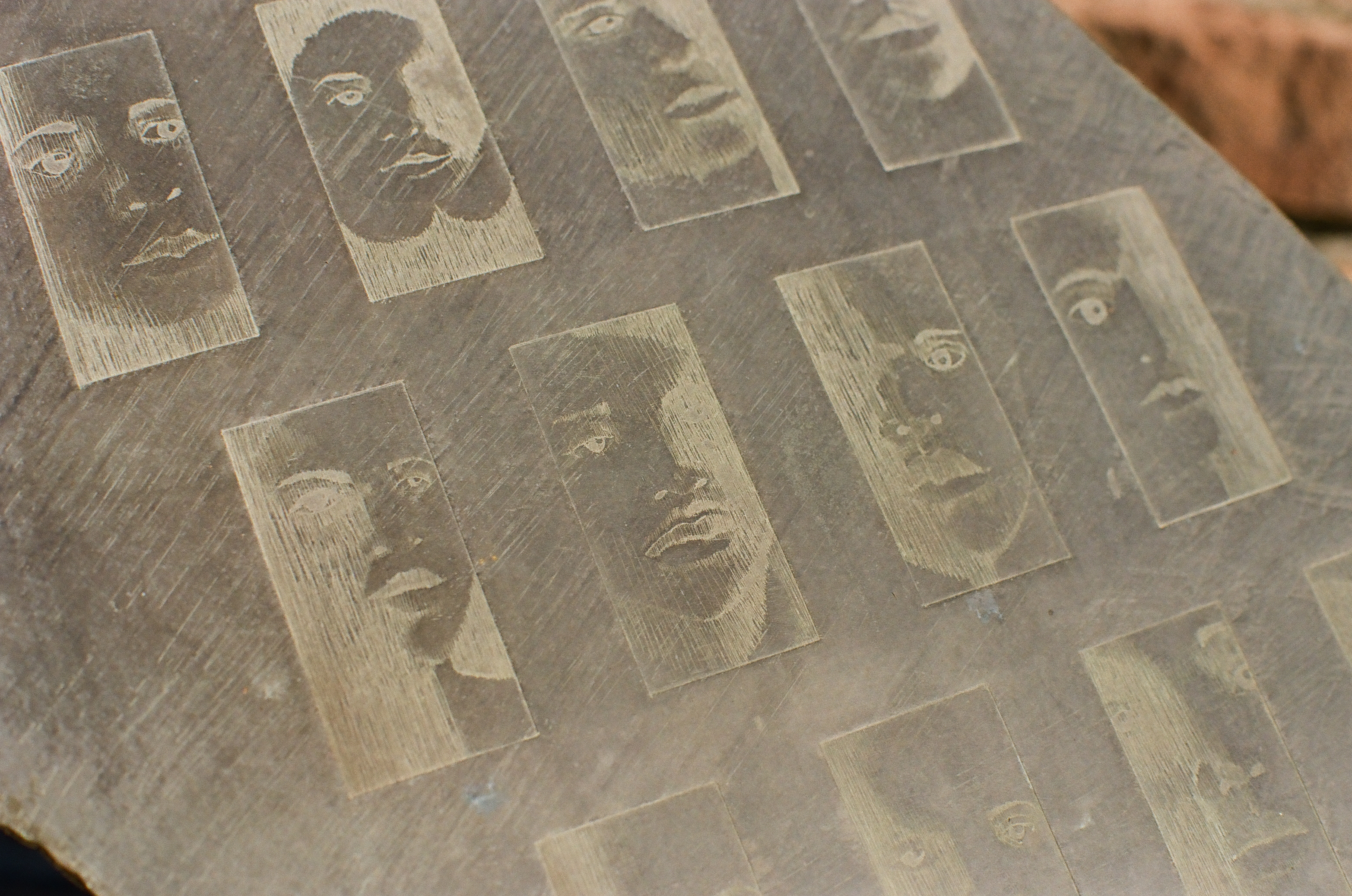 Rows of rectangular panels etched with human faces fill a gray stone slab, with a sliver of reddish brick visible past its upper edge.