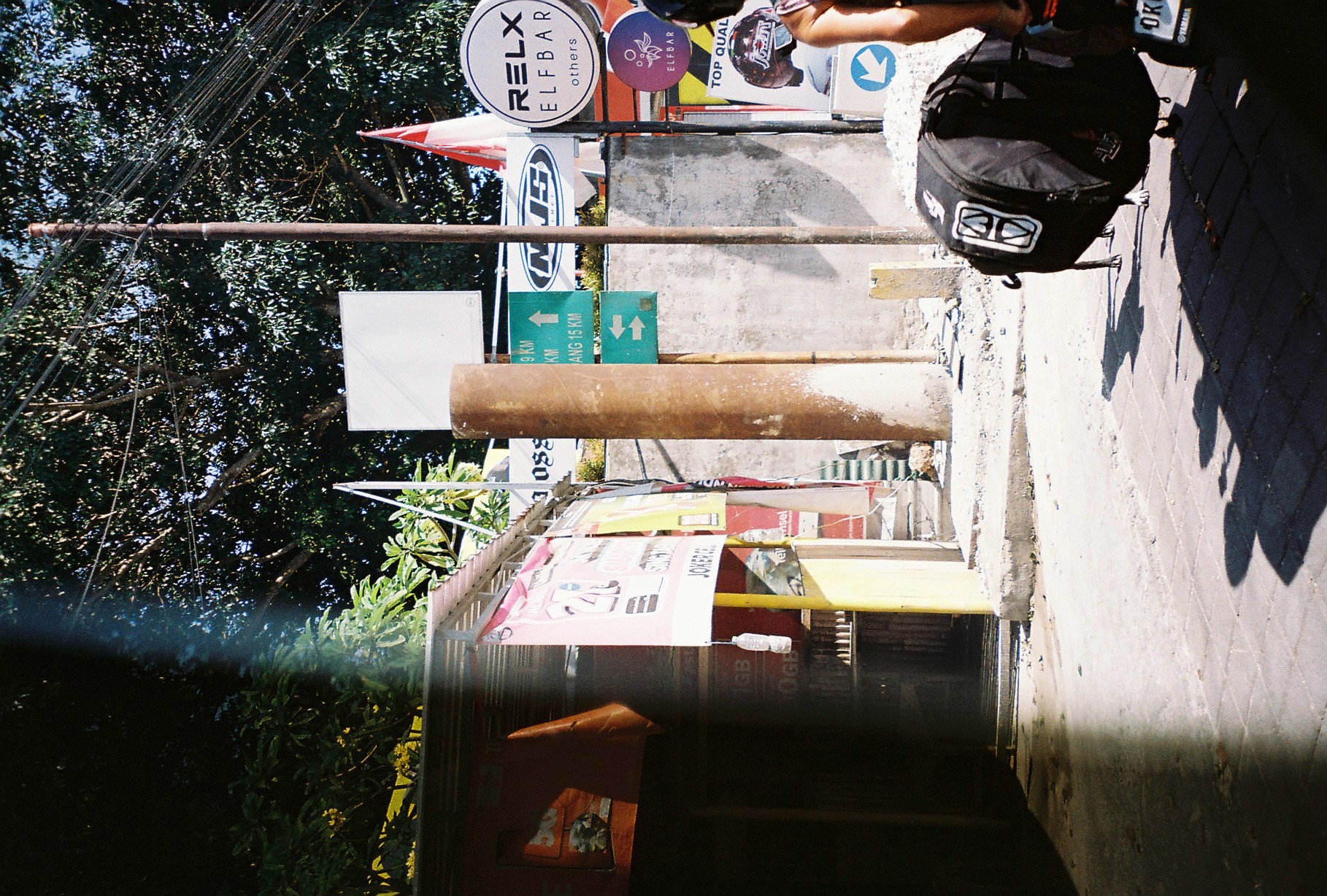 A person's bare arm carries a large black duffel bag into a shaded street corner lined with shop signs and road markers.