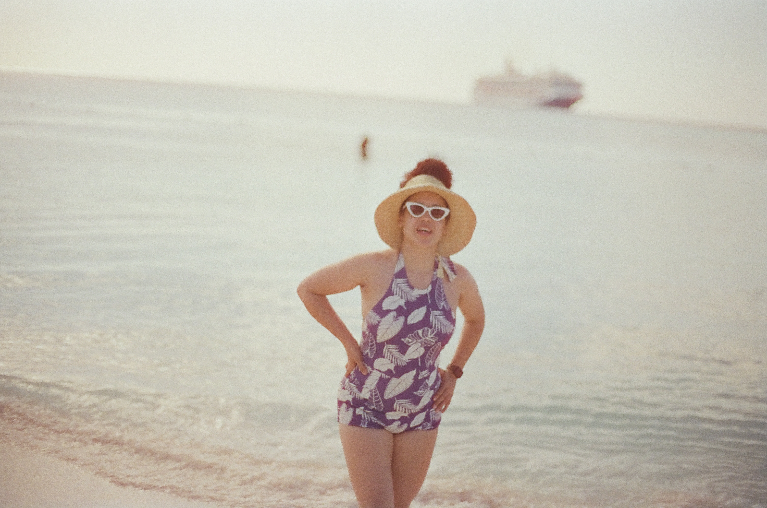 A woman in a straw hat and leaf-print swimsuit poses with hands on her hips at the shoreline, smiling, with a cruise ship blurred on the horizon behind her.