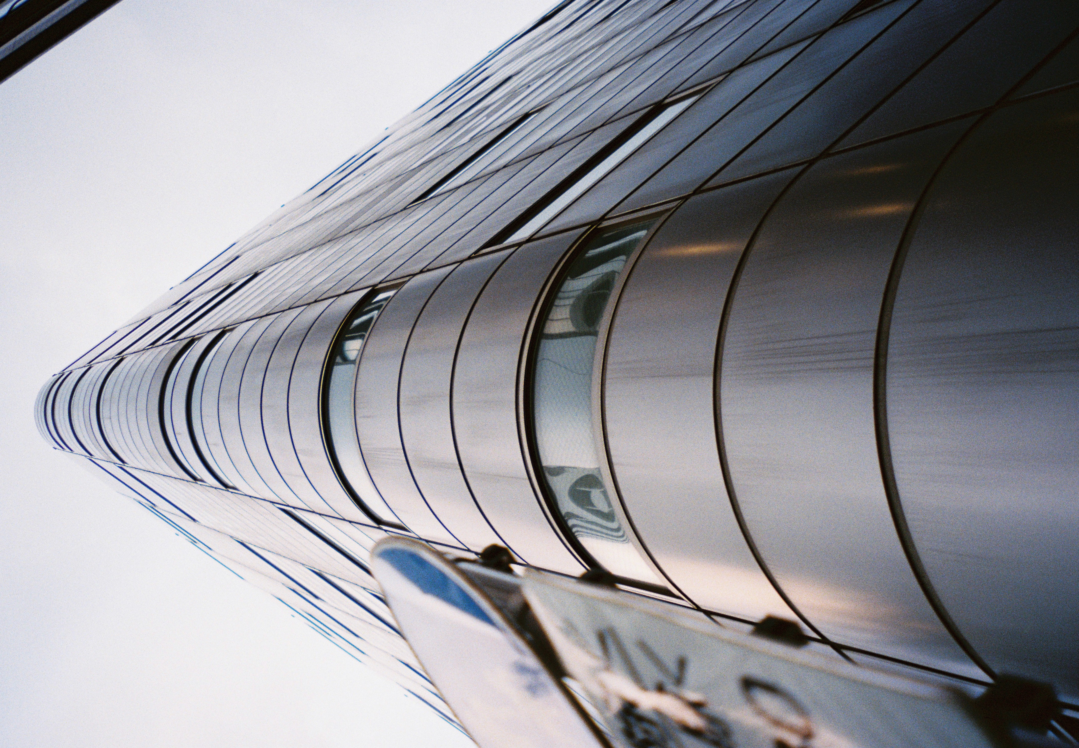 Upward view of the same curved steel building corner, with a blurred street sign intruding at the lower left of the frame.
