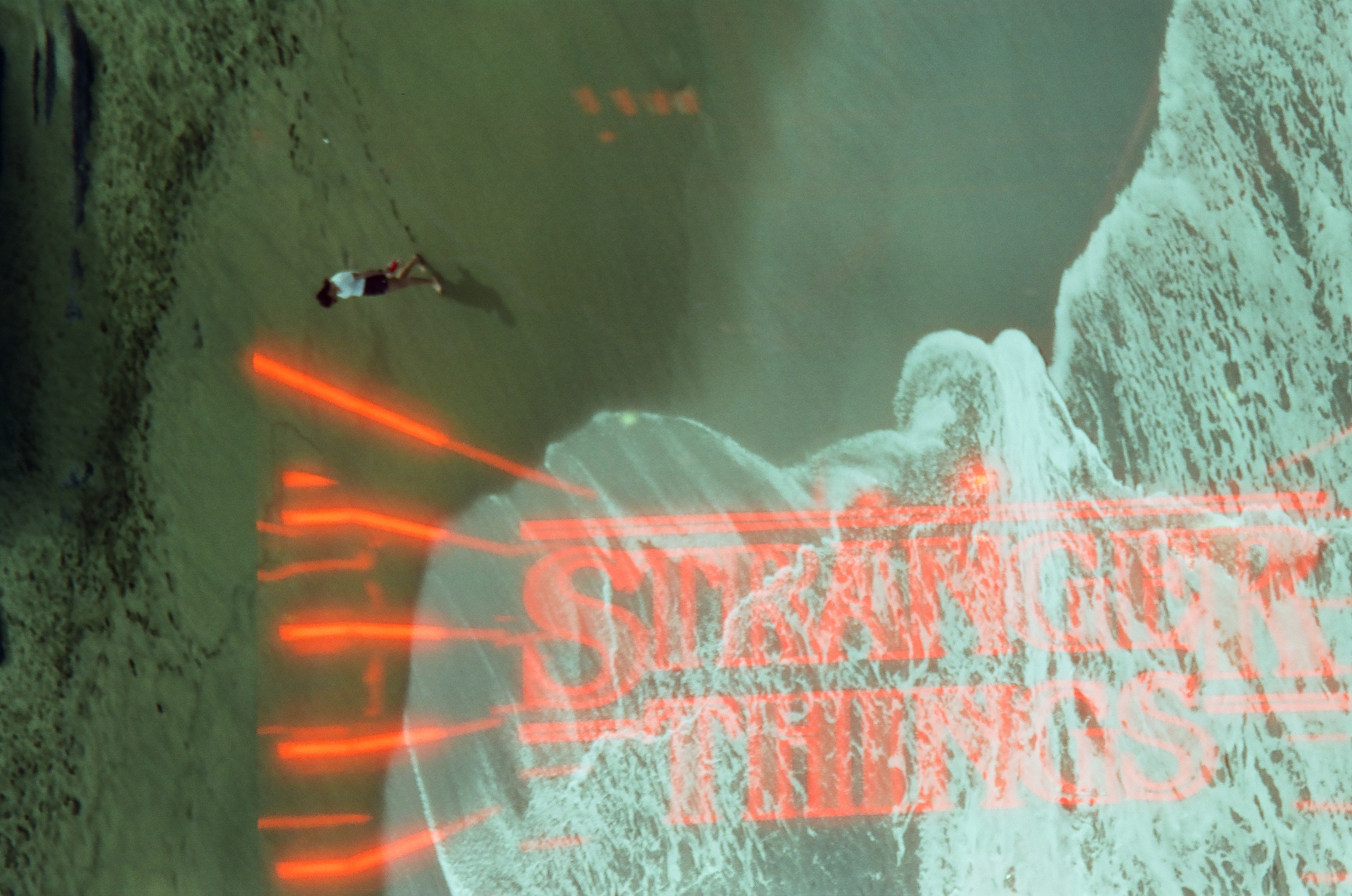 A lone figure in a white top walks across open sand, dwarfed beneath large translucent red neon lettering overlaid on the scene.