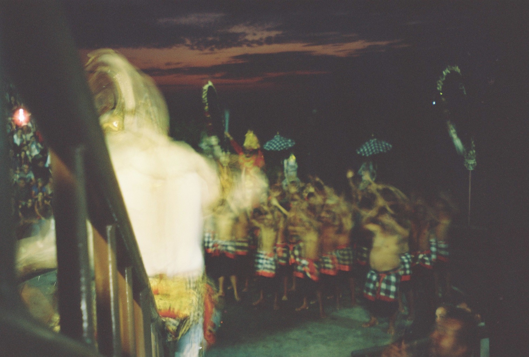 A heavily blurred pale figure fills the foreground before rows of bare-shouldered people in checked cloth beneath a dark red-streaked sky.