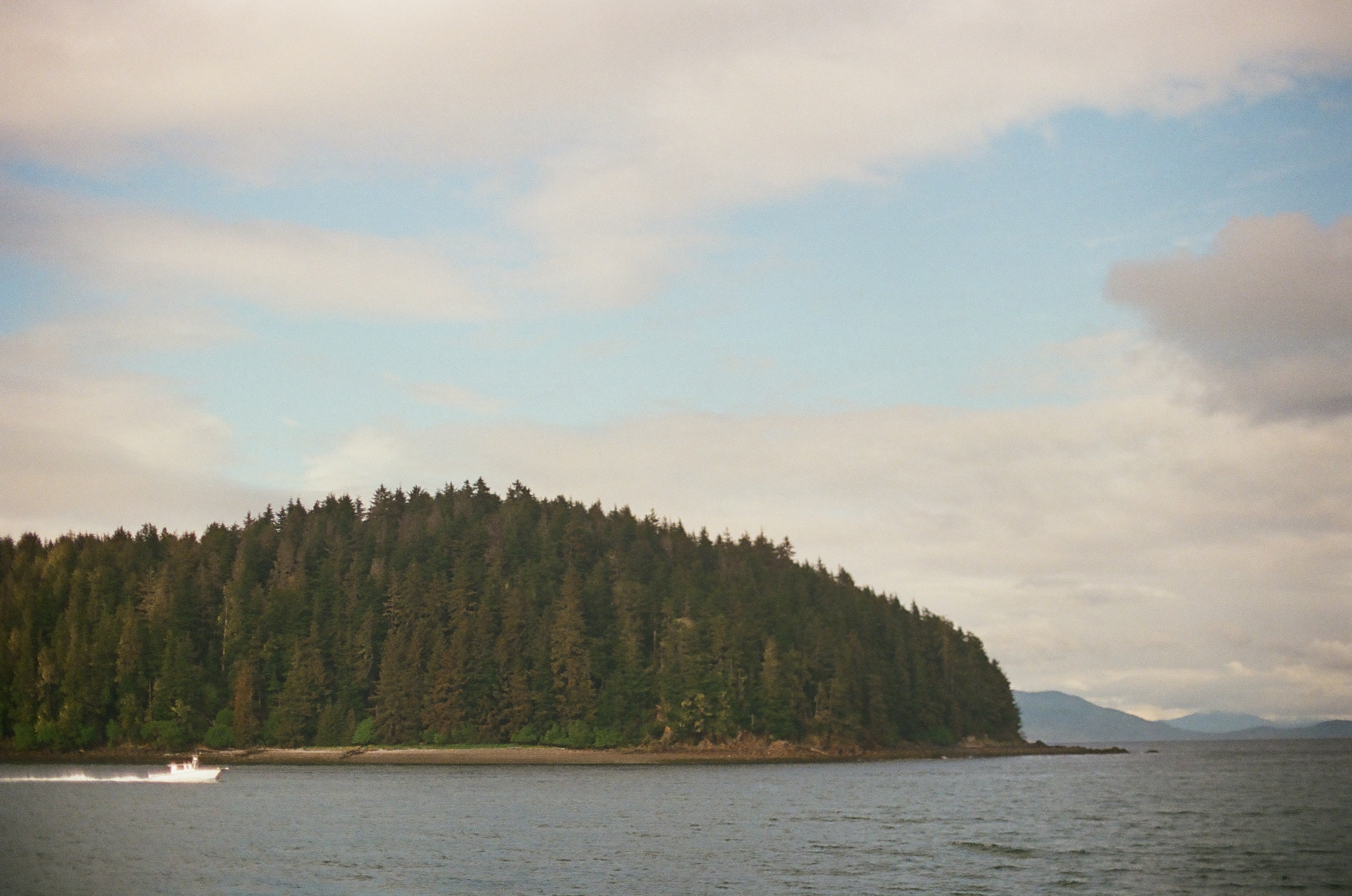 A long forested island stretches across still water beneath gray clouds and a pale blue opening.