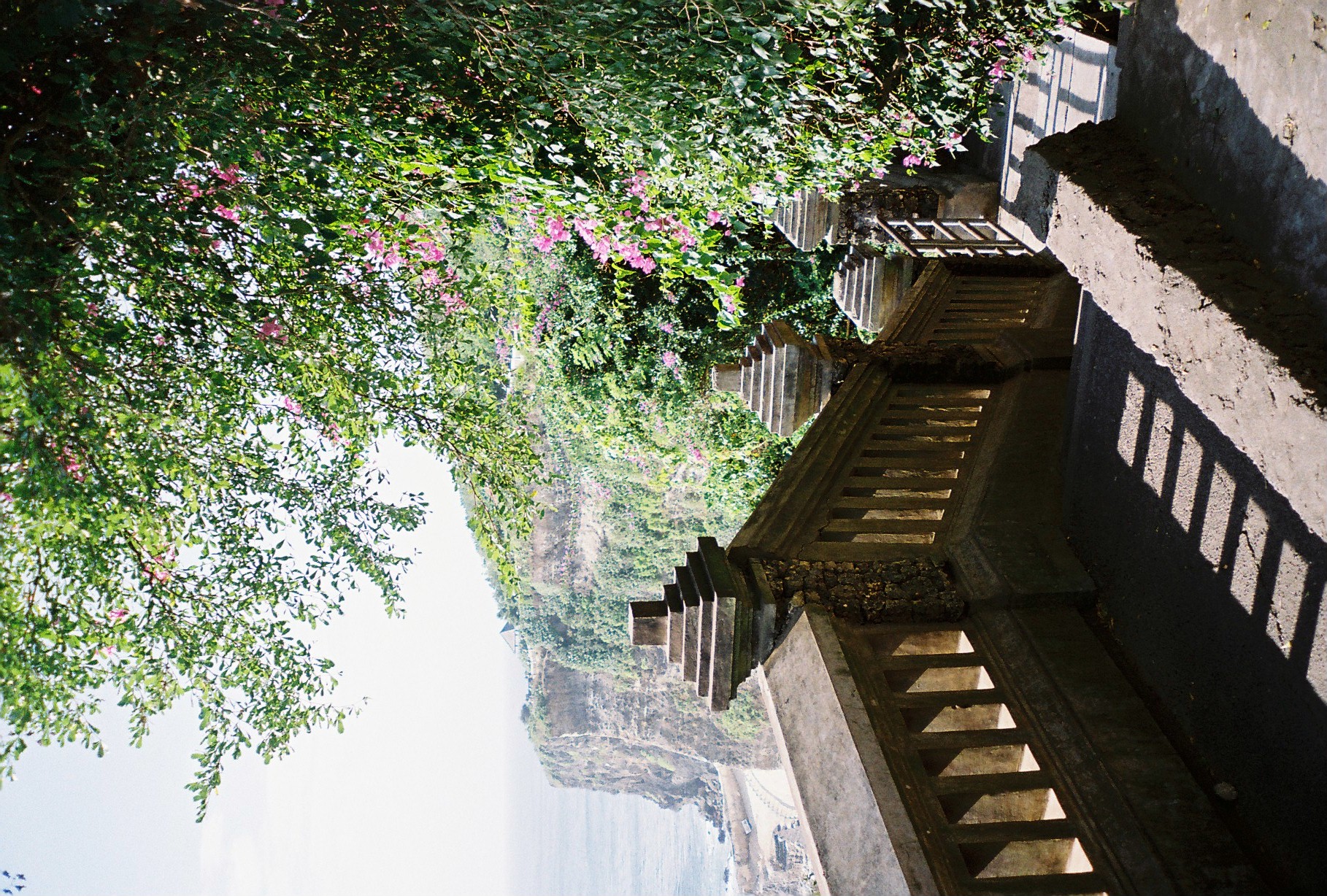 A stone staircase lined with square balustrade posts descends beneath flowering bougainvillea and tree branches toward a hazy strip of ocean below.