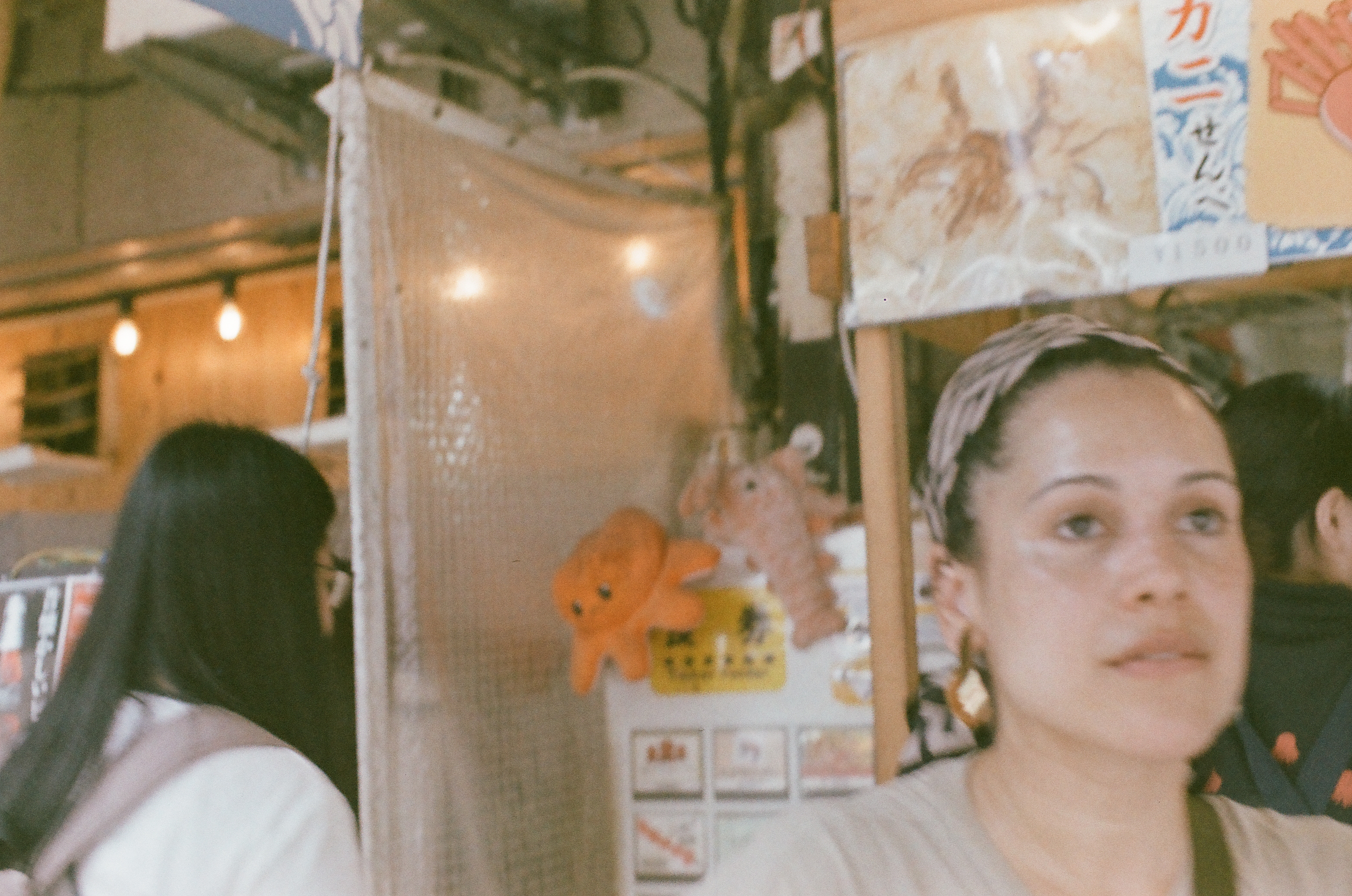 Gold-toned hoop earrings catch the light as a woman glances toward the camera beside a stall shelf holding a plush orange octopus toy.