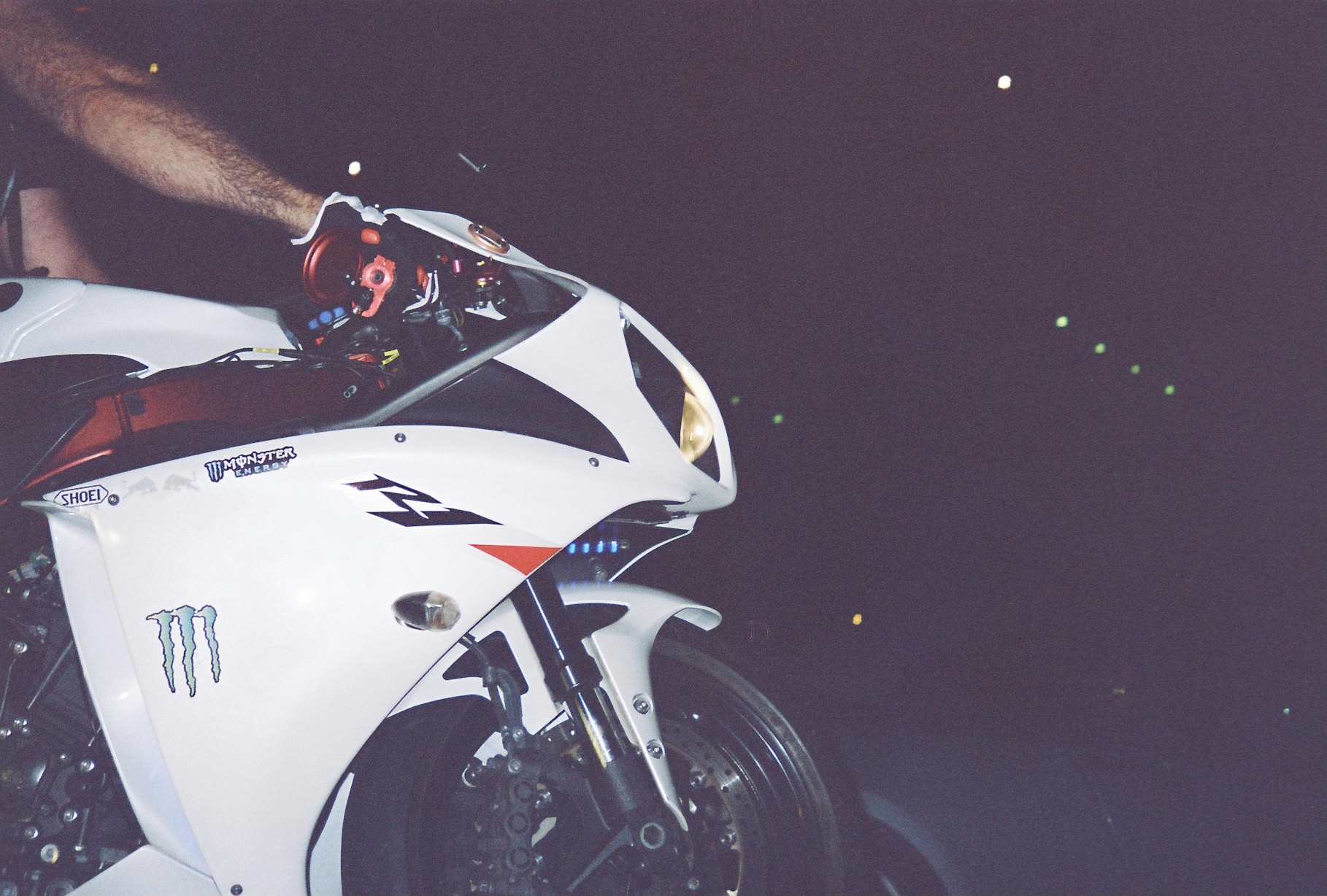 A white sport motorcycle fills the left half of the frame while a bare arm reaches down to a gloved hand on its handlebar against darkness.
