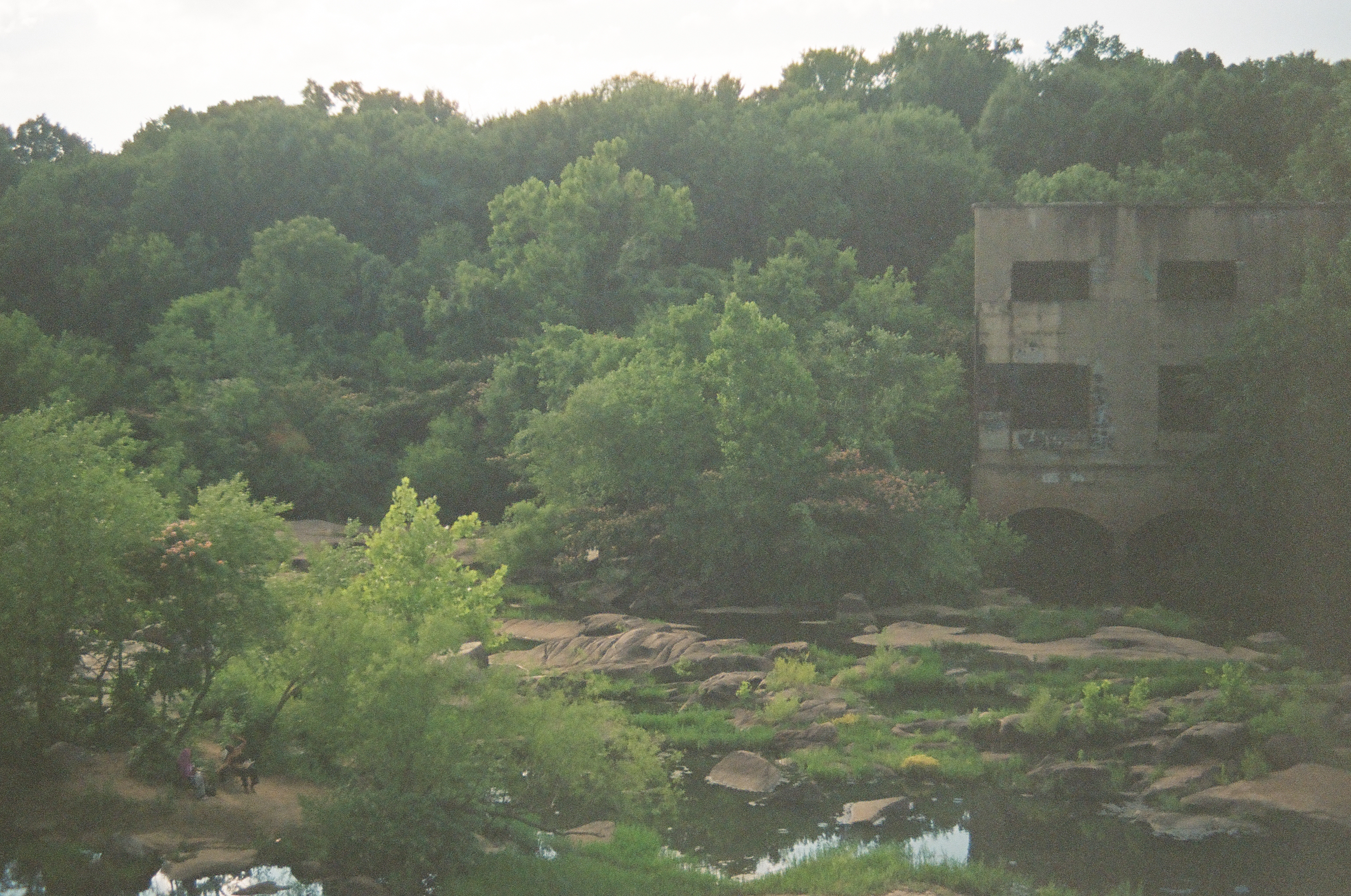 A multi-story concrete building with dark window openings and graffiti stands above a rocky riverbed, with two small figures visible on a path near shallow pools below.