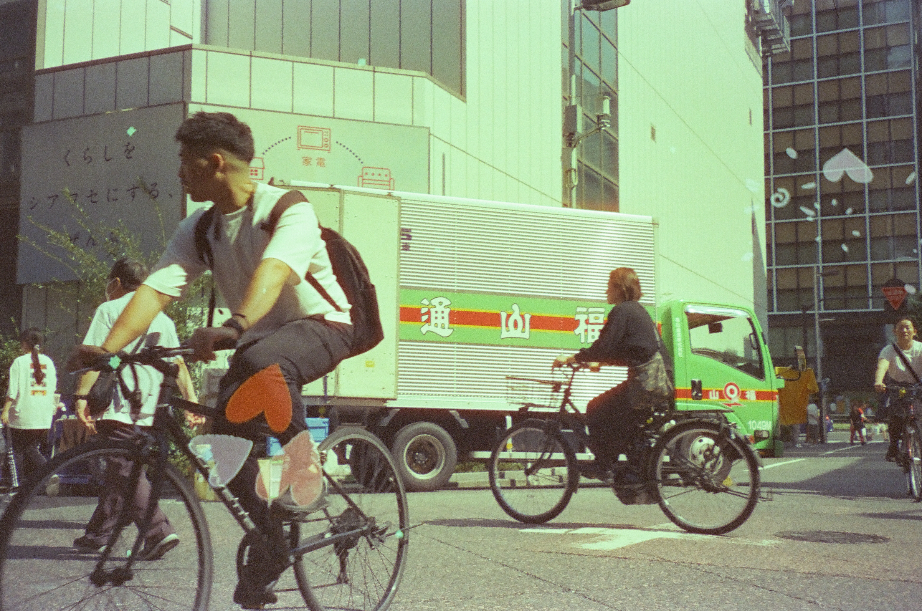 Two cyclists ride past a green delivery truck marked with large Japanese characters, one wearing a white shirt and backpack, near a building with printed Japanese text.