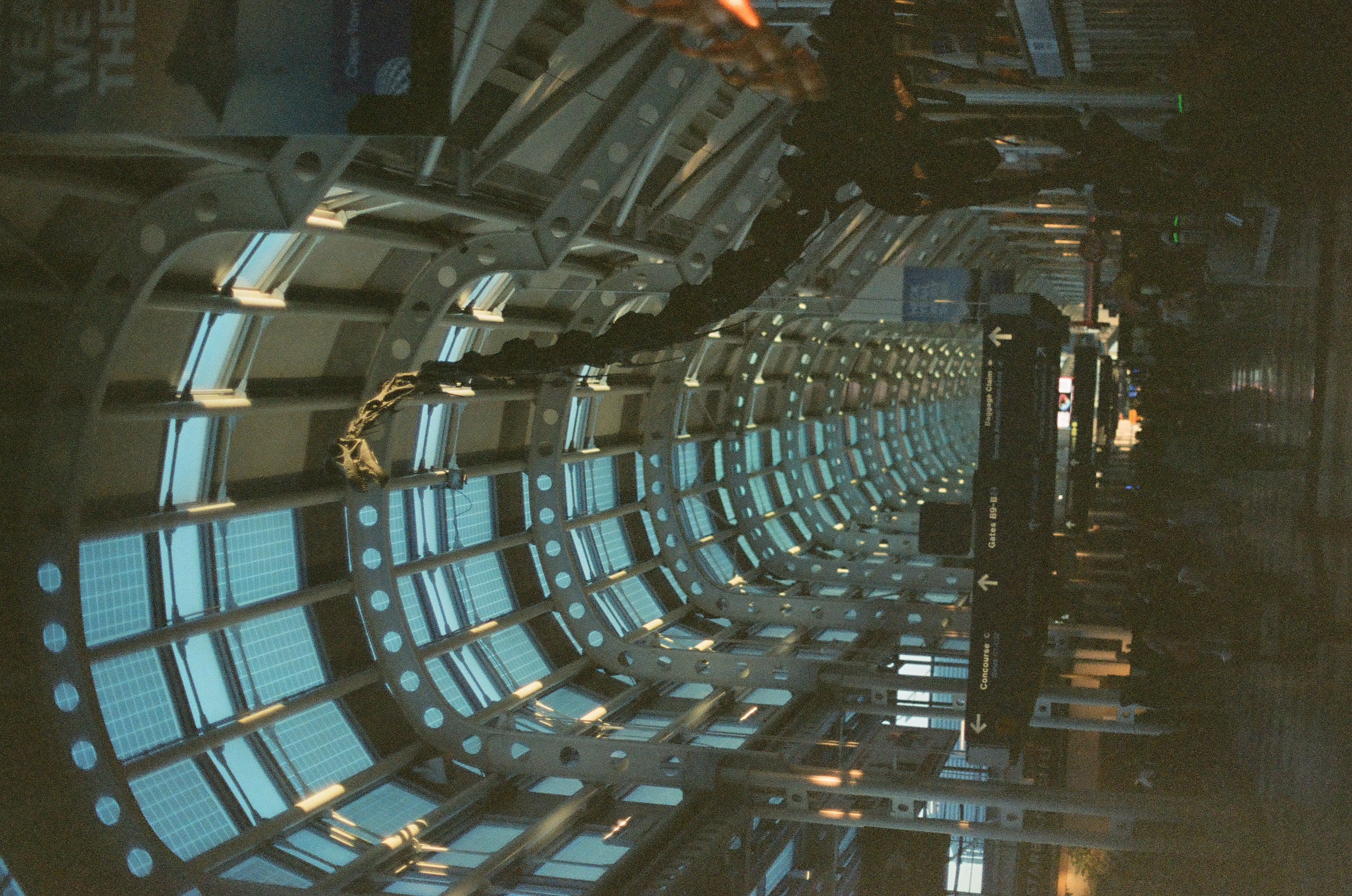 A long airport concourse of curved glass and steel ribs extends toward distant travelers under dim green light.