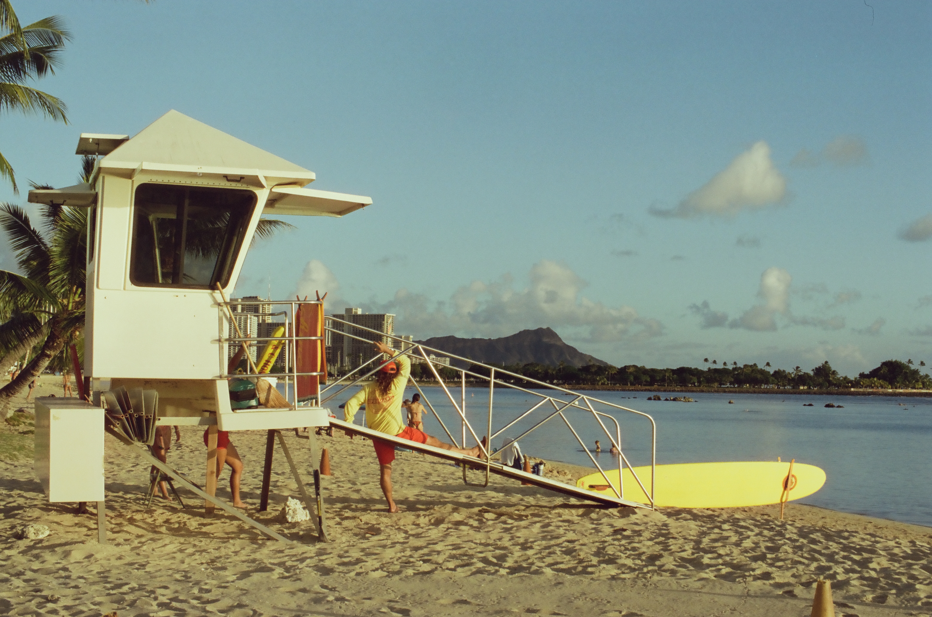 Sand fills the foreground beneath the lifeguard tower and ramp, an orange cone and the yellow board sitting near the water's edge.