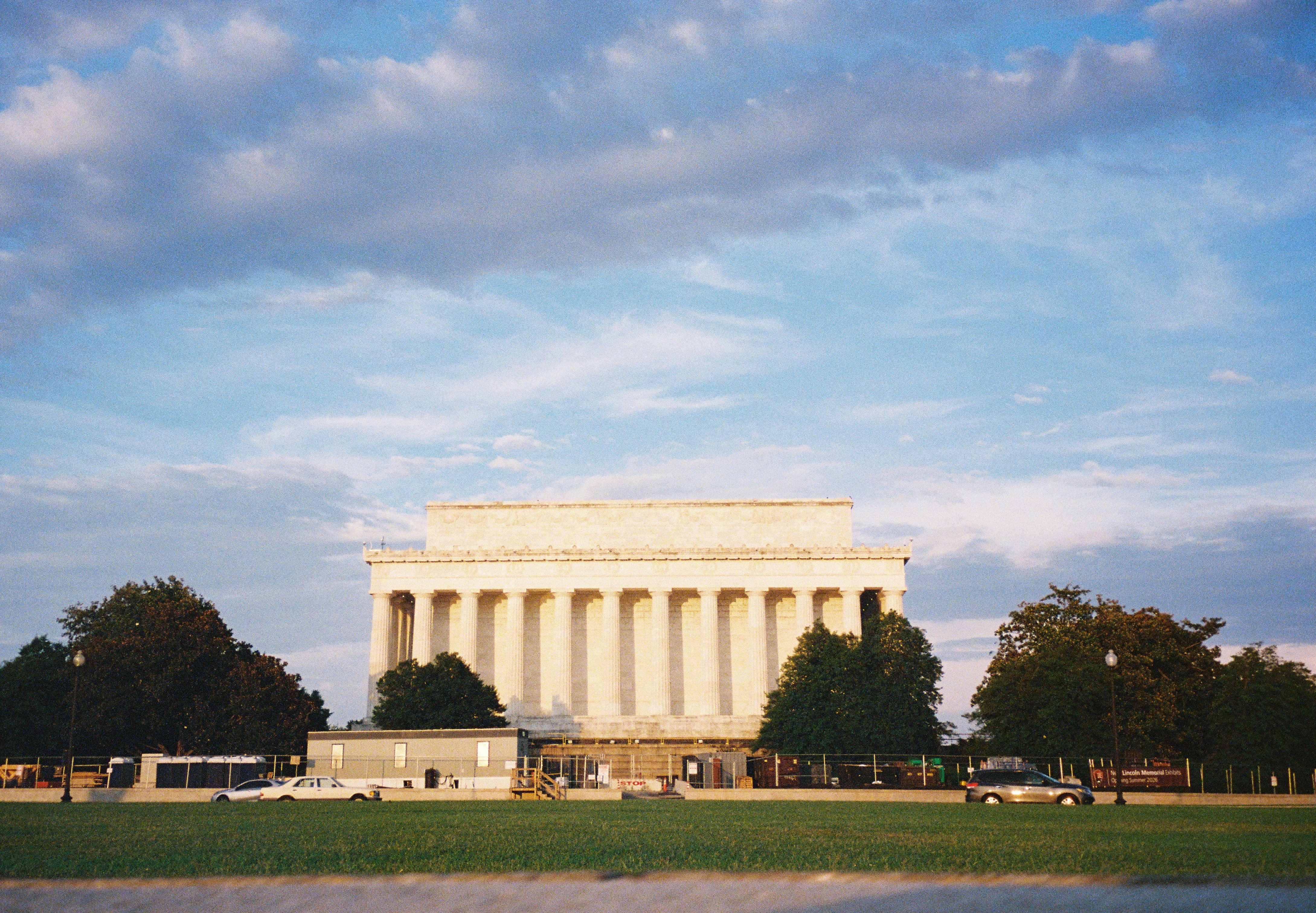 A pale columned memorial sits across a wide green lawn beneath a cloud-streaked blue sky.
