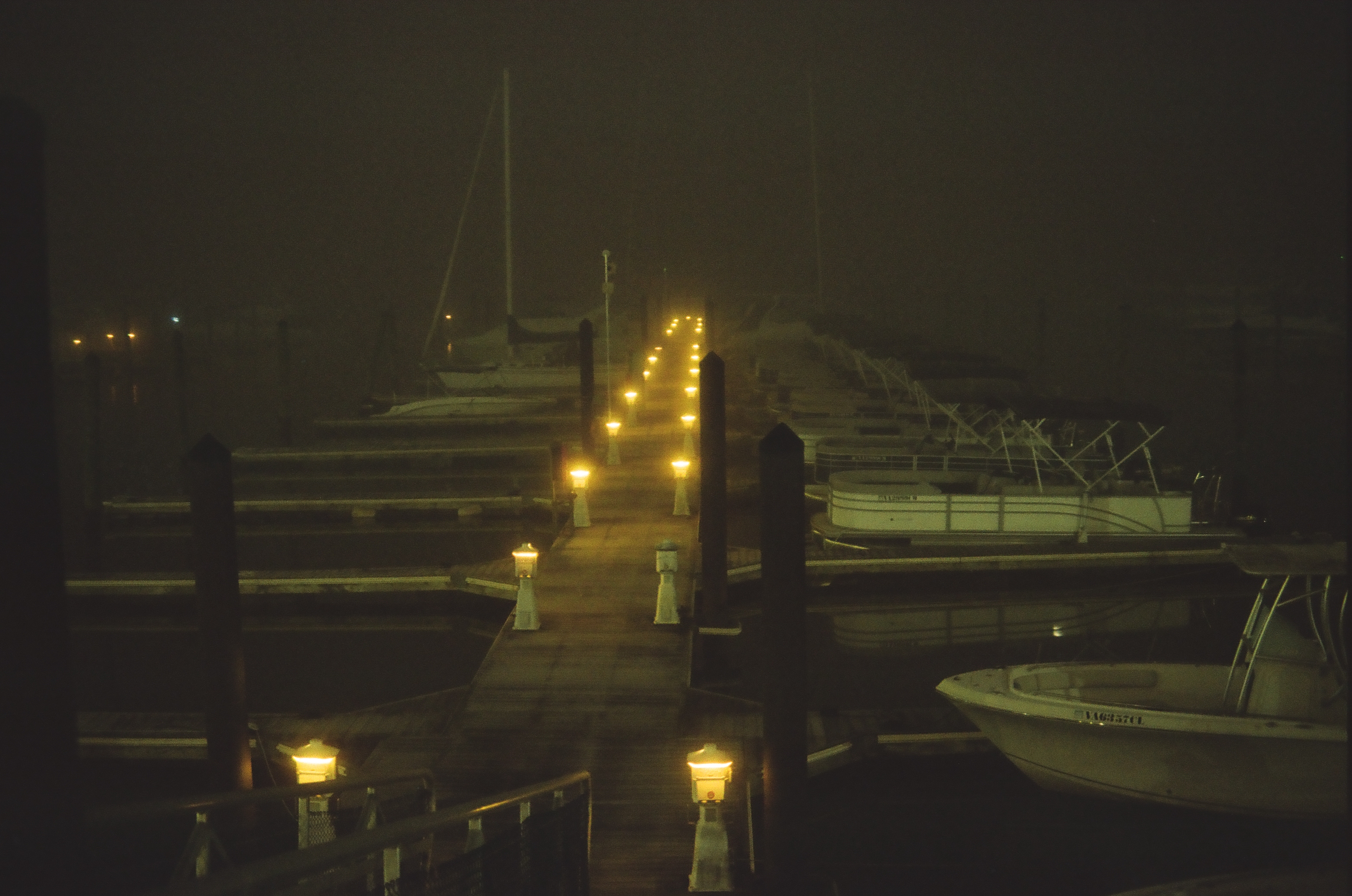 A long wooden dock lined with small lamps recedes into fog between moored sailboats and covered powerboats at night.