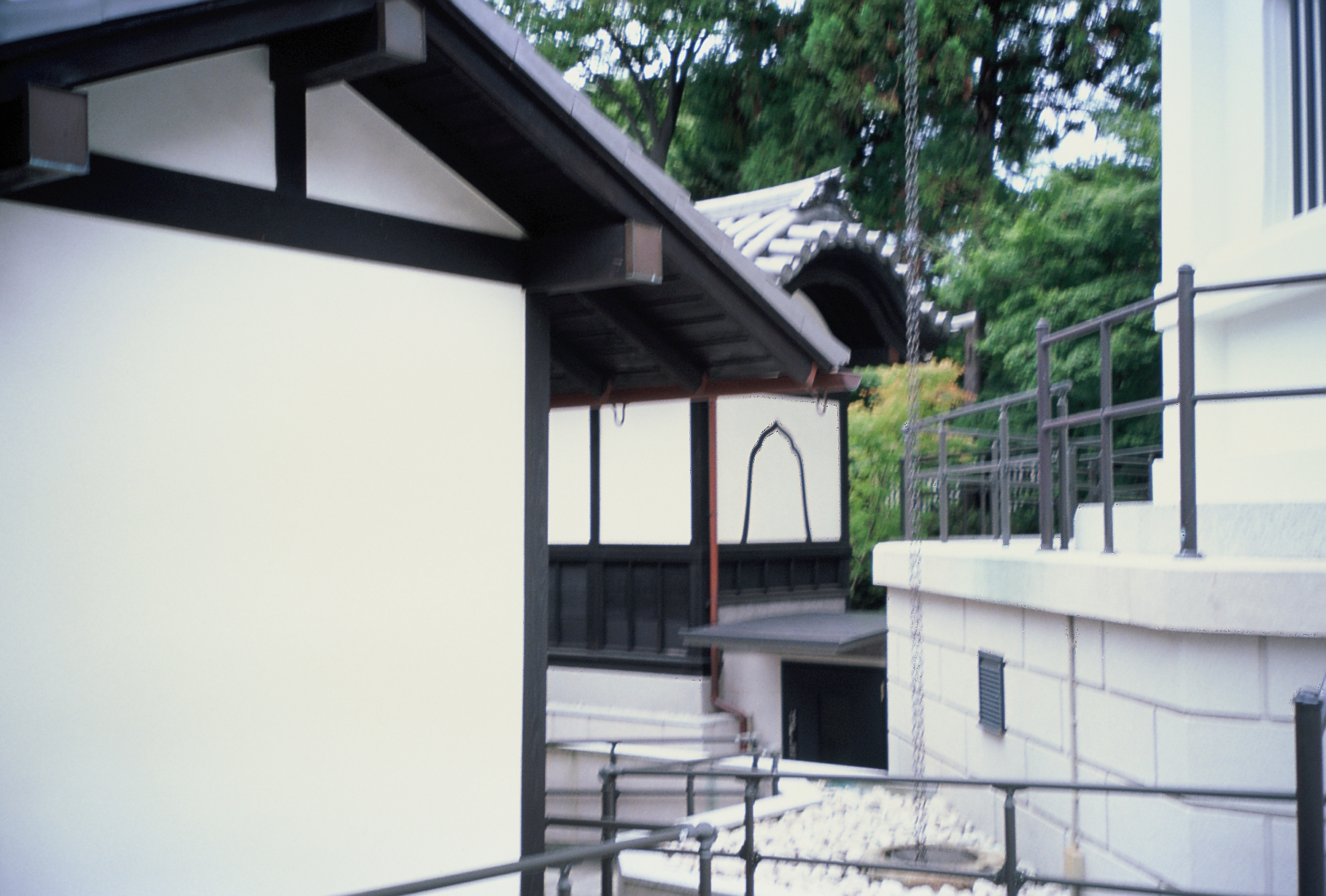 Dark timber eaves of a plain building frame a curved temple roof glimpsed through trees, with a white balcony and railing at lower right.
