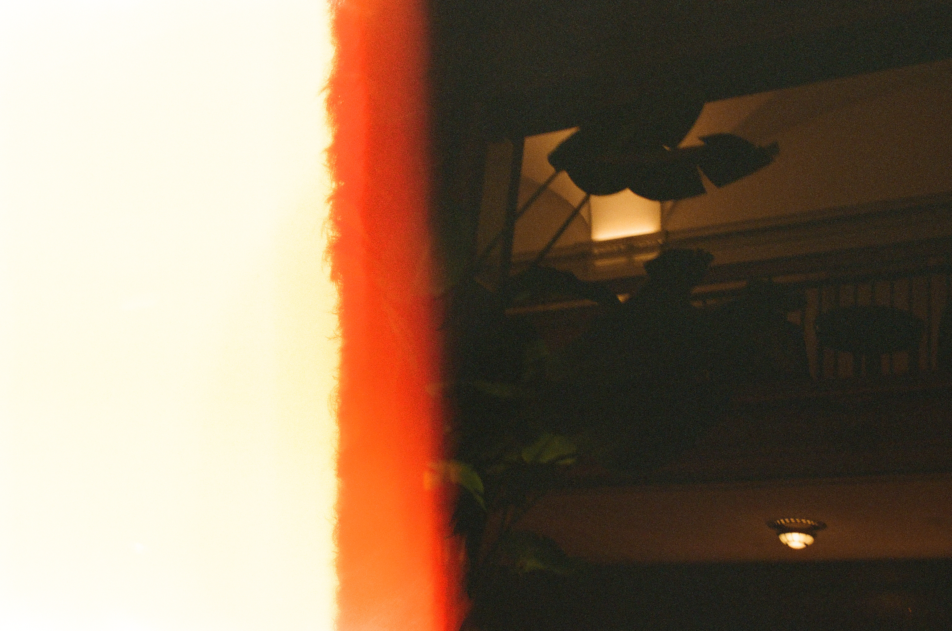 A bright orange vertical film-edge streak cuts across a dim room, where a dark ceiling fixture and a small round light are barely visible.