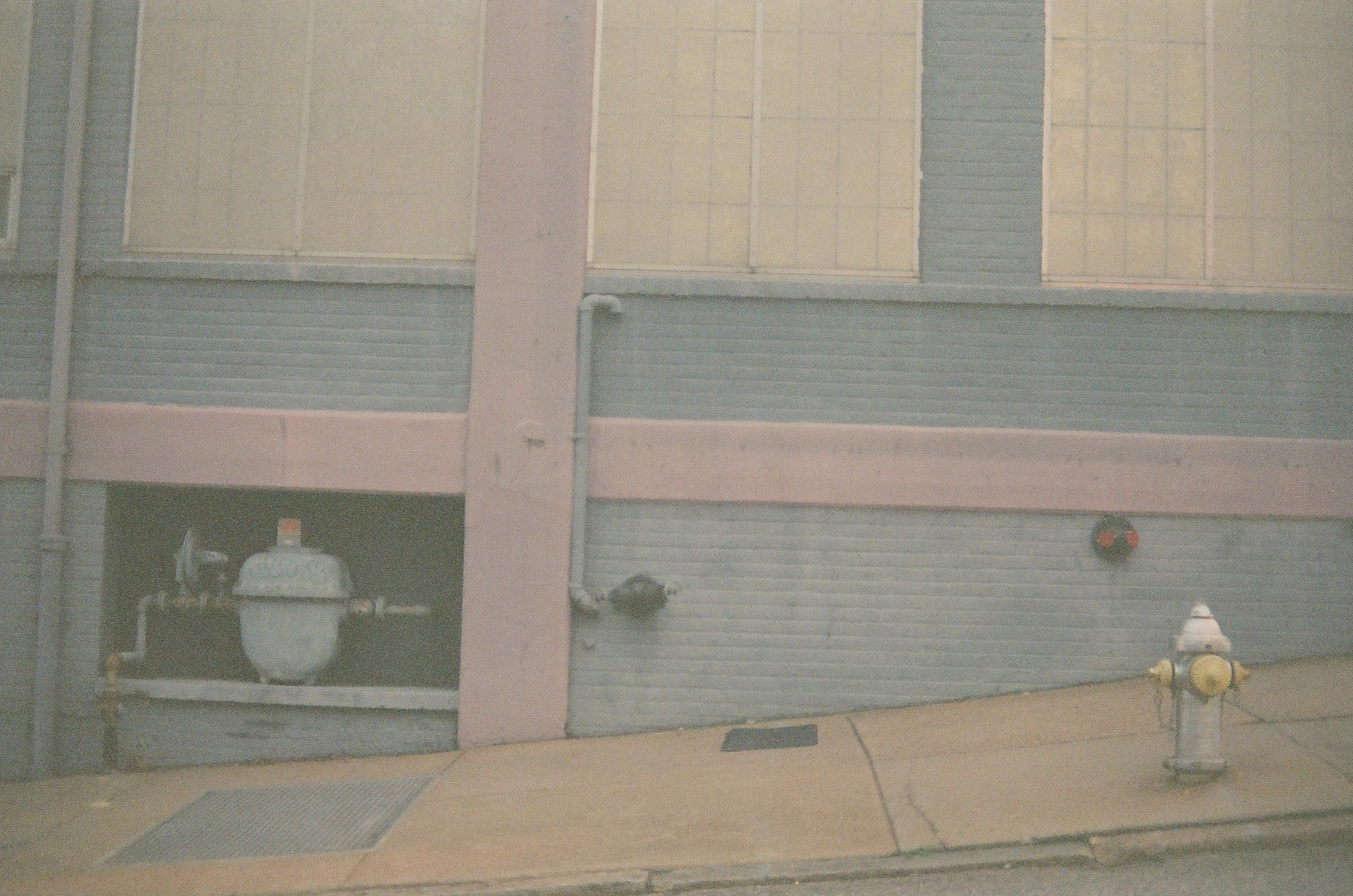 A blue-gray and pink industrial wall holds a recessed gas meter, exposed pipes, and a white fire hydrant on a sloped sidewalk.