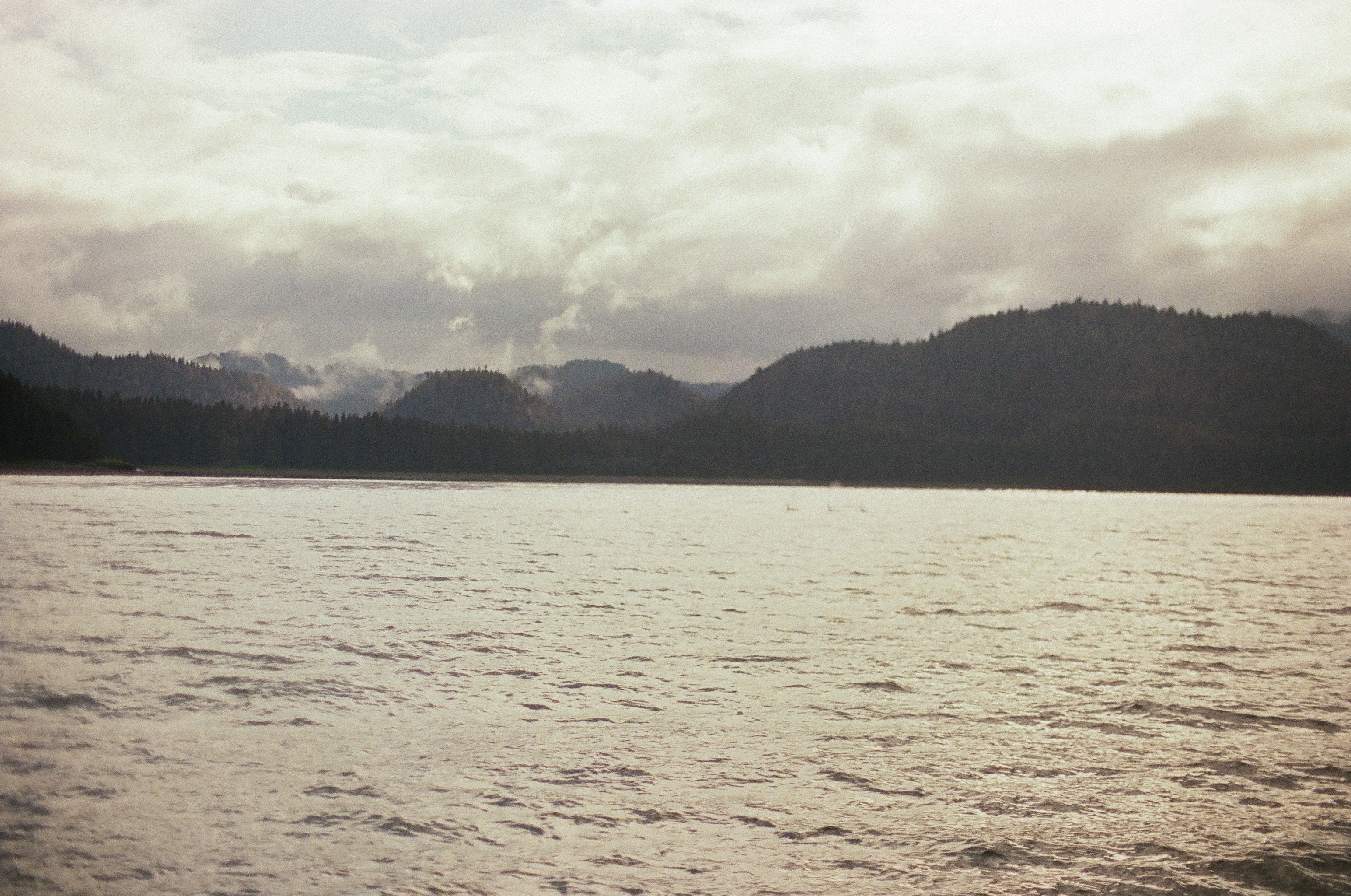 Pale rippled water leads toward overlapping forested ridges and distant mountains beneath low layered clouds.