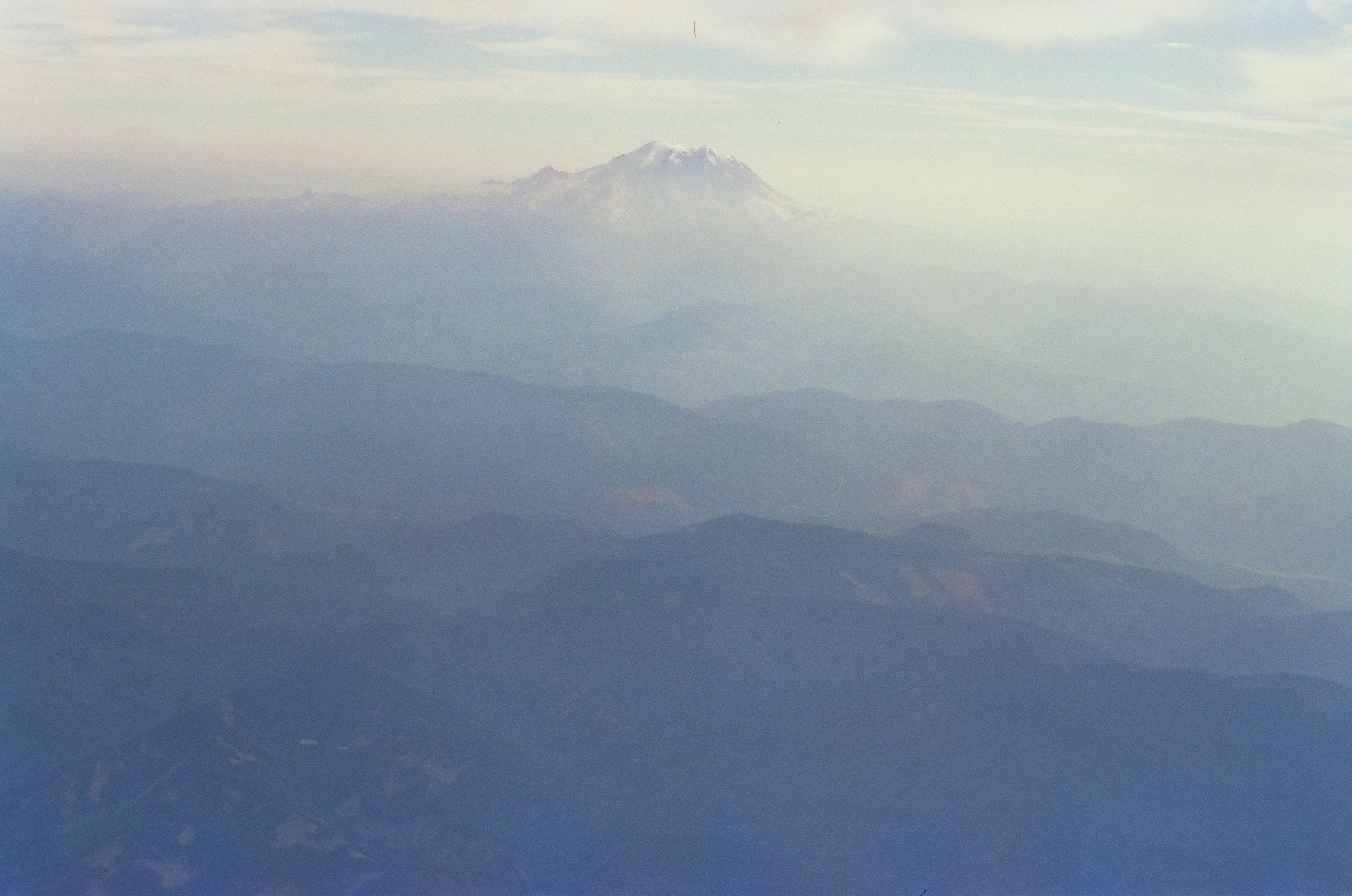A snow-capped peak sits near the top center of the frame above tan-toned foothills and layered blue ridgelines beneath a warm-hazed sky.