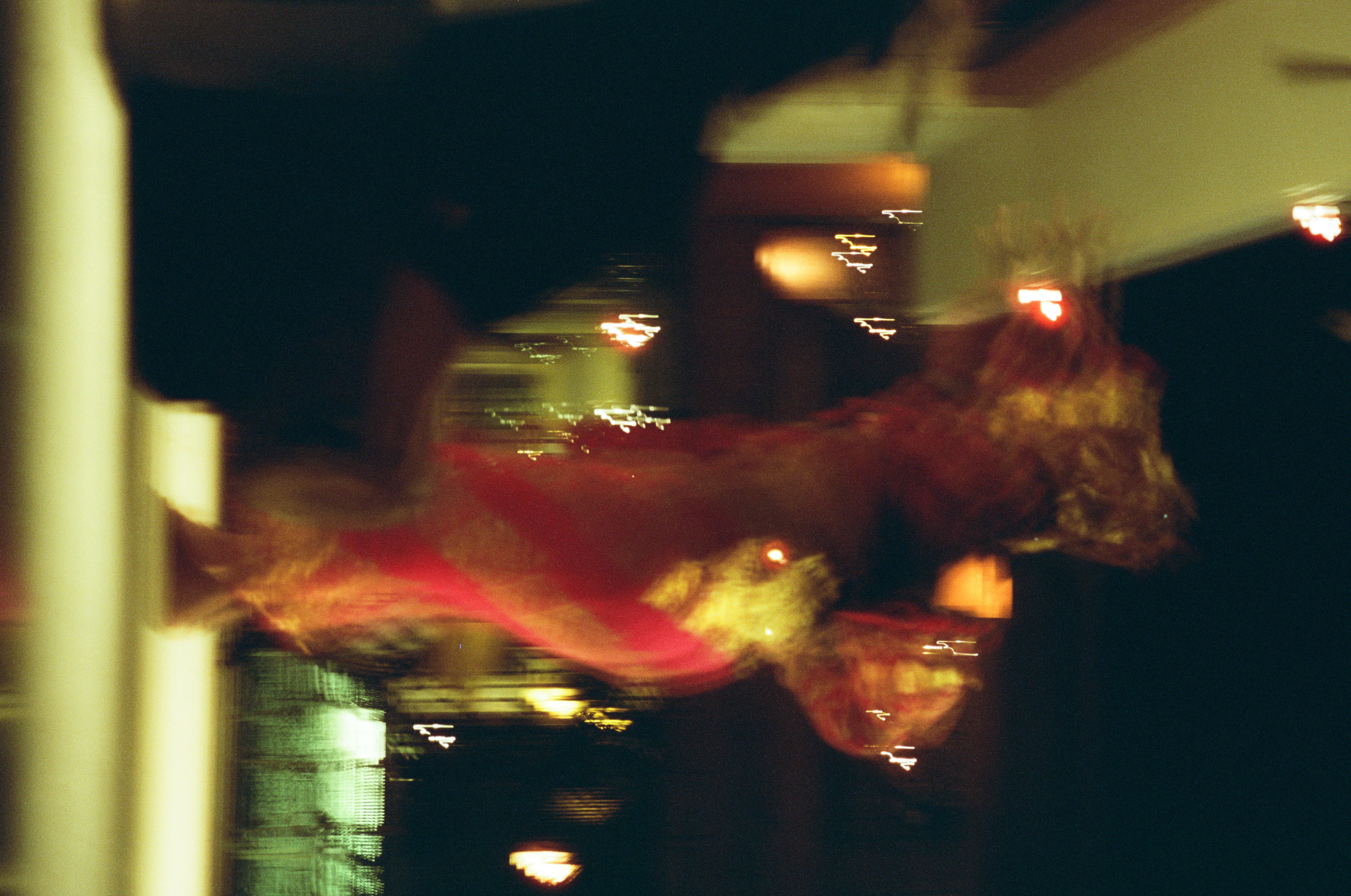 A blurred red-and-gold figure with a large gold head adornment lifts one arm overhead while the other bends outward amid streaked lights.