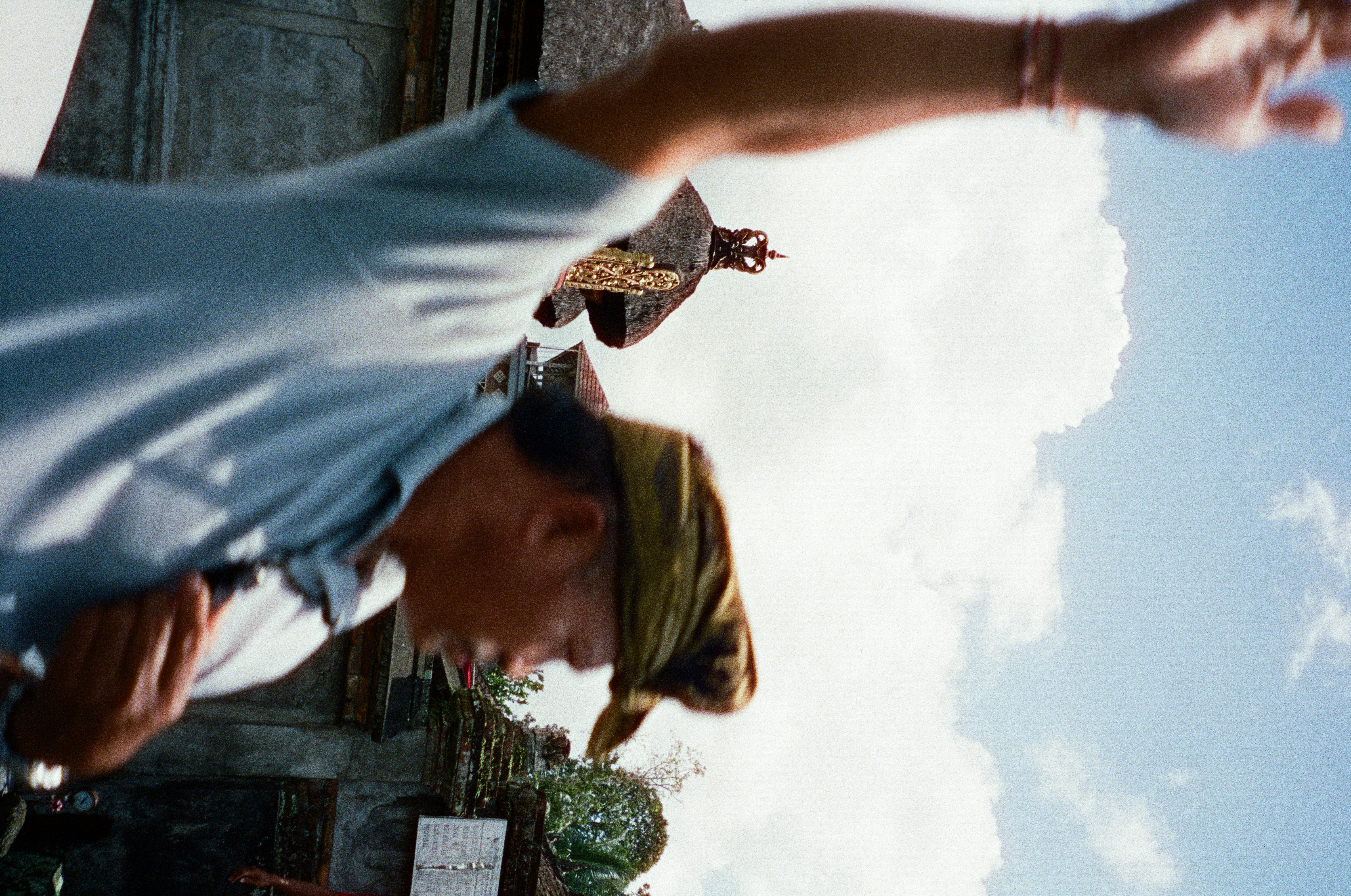 A blurred adult in a patterned head wrap raises one arm before white clouds, walls, and an ornamented thatched roof.