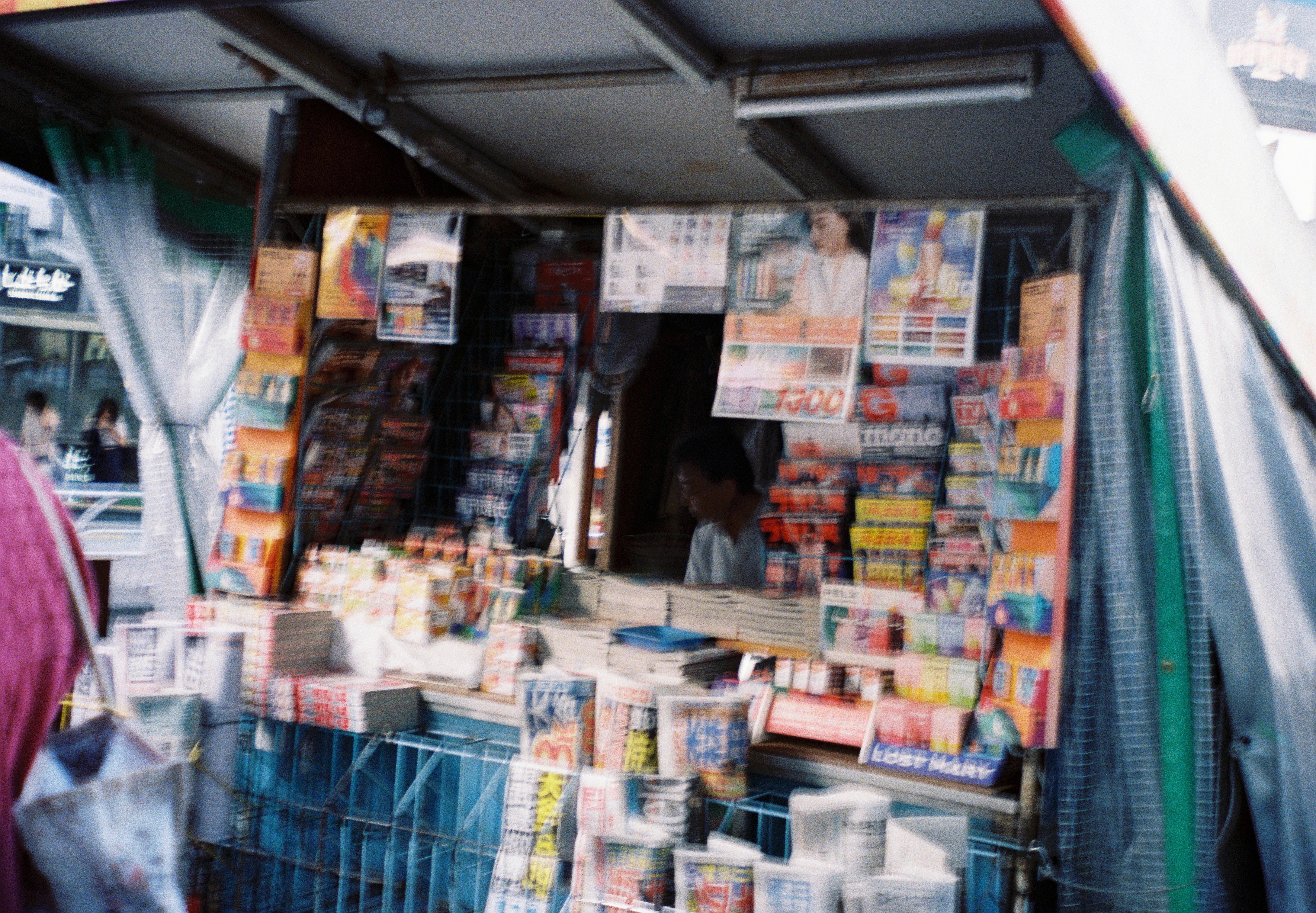 Racks of magazines in plastic sleeves fill a newsstand stall rendered in soft focus, with a person faintly visible in the dim interior.
