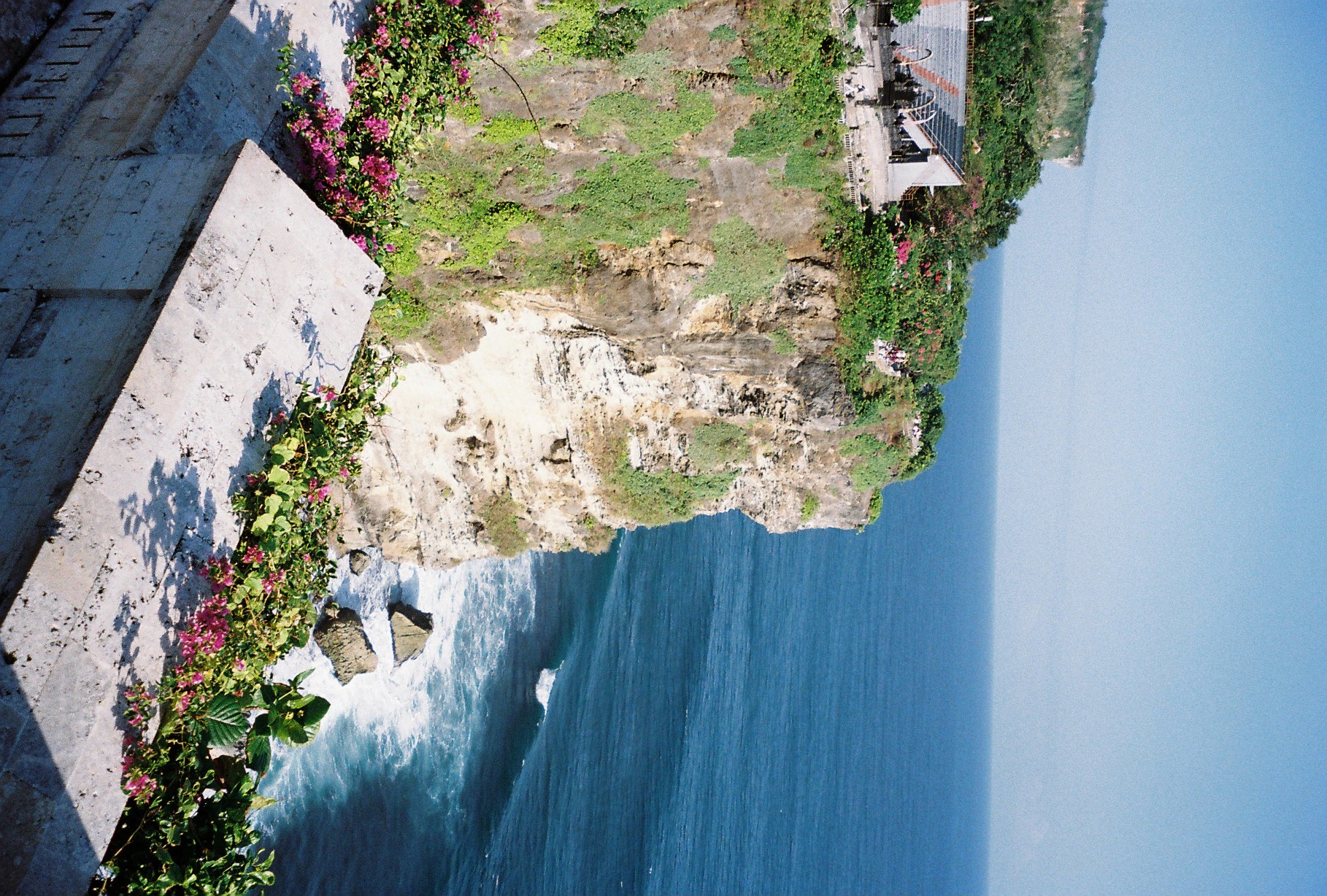 A rocky, scrub-covered headland juts into blue ocean, with white surf breaking on rocks below and a railed building visible among trees at its crest.