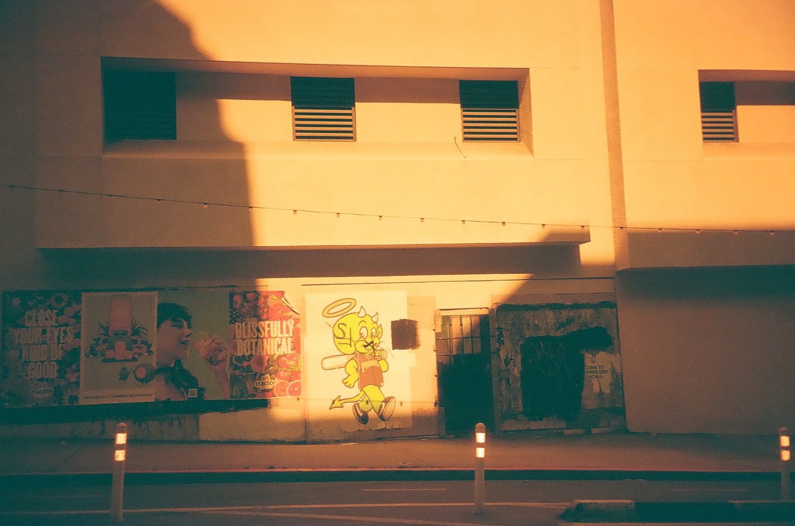 Posters and a painted cartoon devil-angel figure line a sunlit wall beneath a string of small lights and a sharp diagonal shadow.