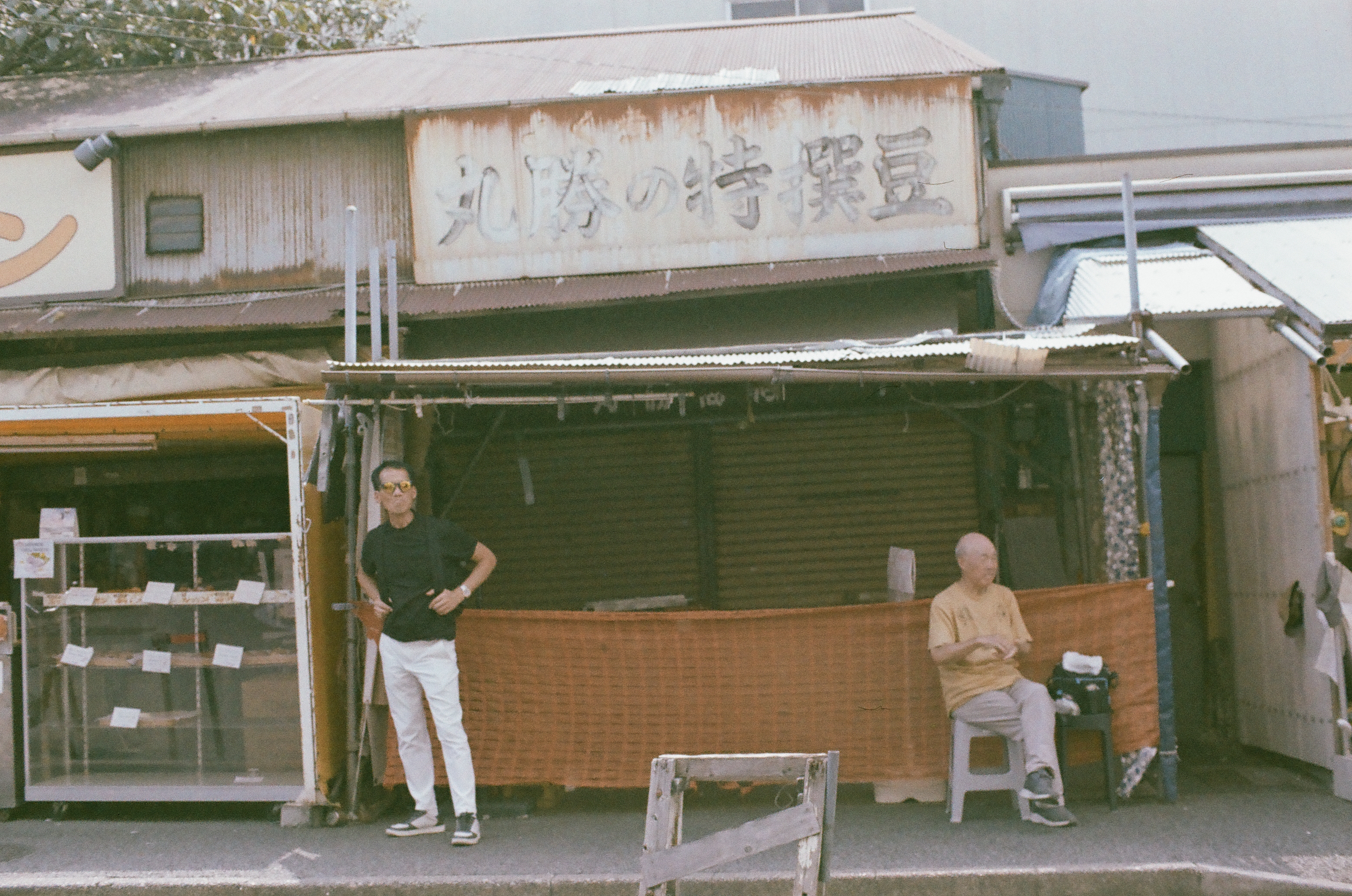 One man stands in sunglasses and white pants while another sits on a stool behind a woven orange screen outside a closed shutter.