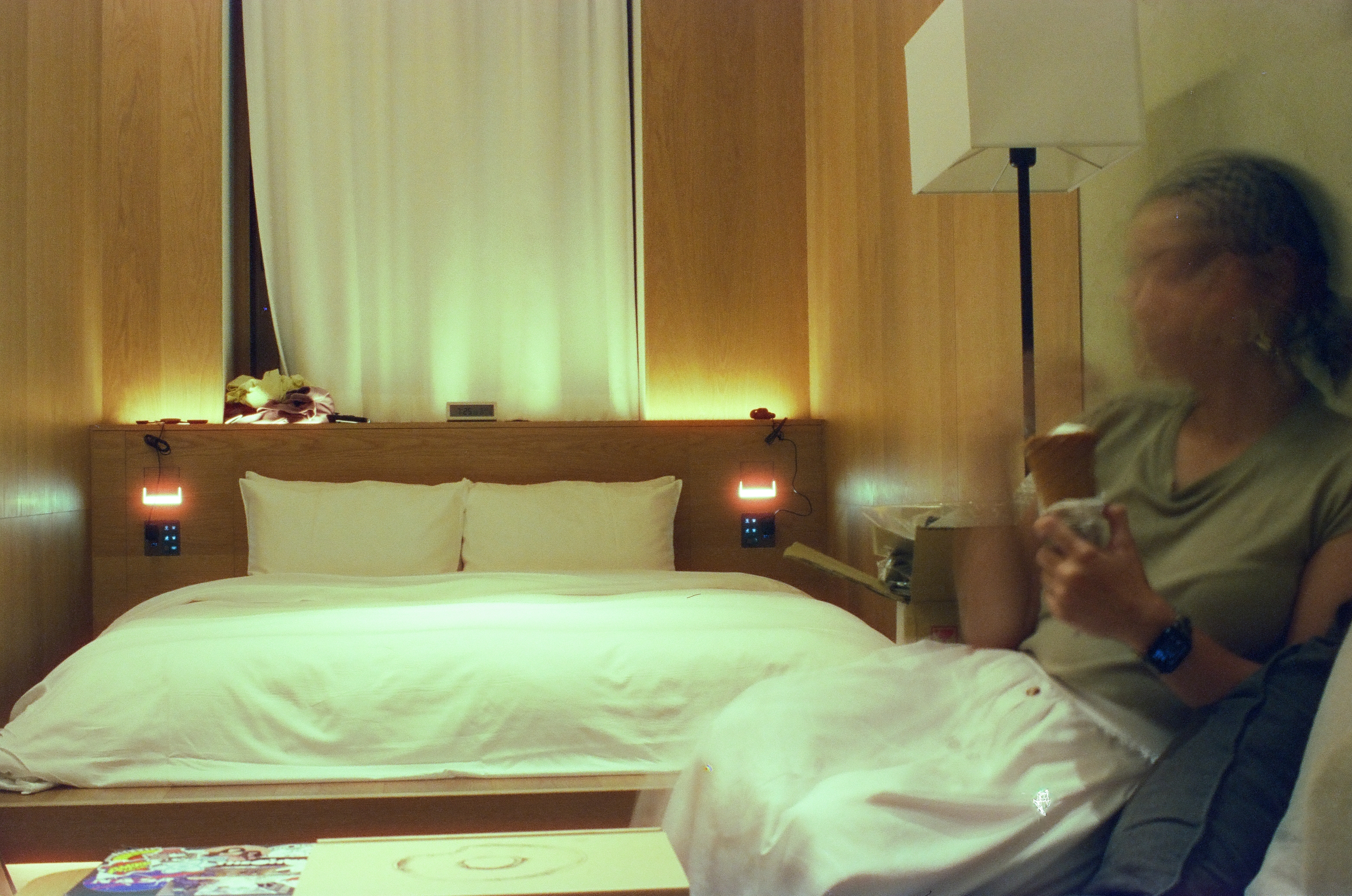 A wood-paneled room with a white bed, two pillows, a backlit white curtain, two small wall lights, and a motion-blurred seated person holding a brown cone-shaped item at right.