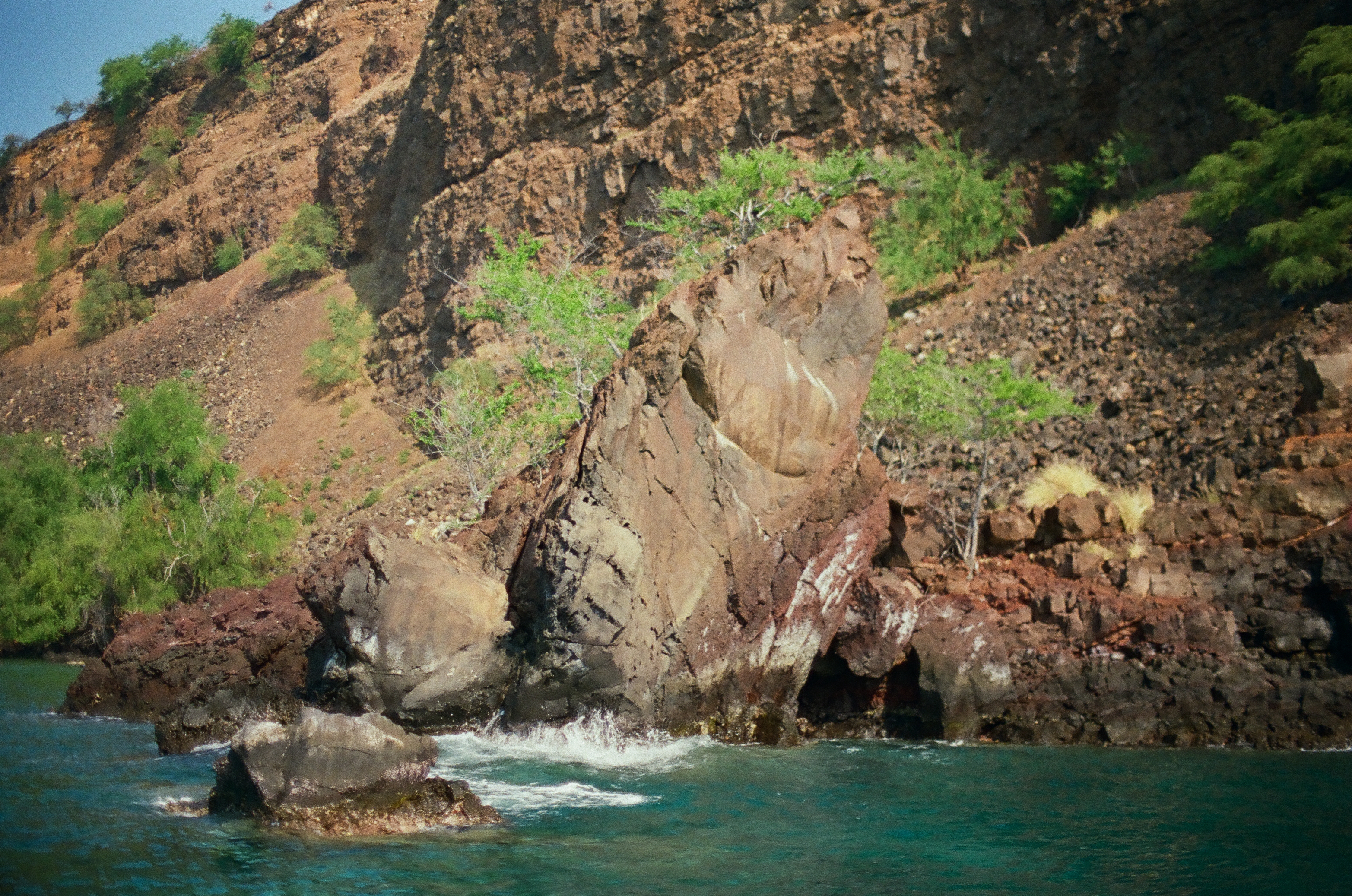 Clear turquoise water breaks against steep red-brown cliffs dotted with green shrubs and dark volcanic rock.