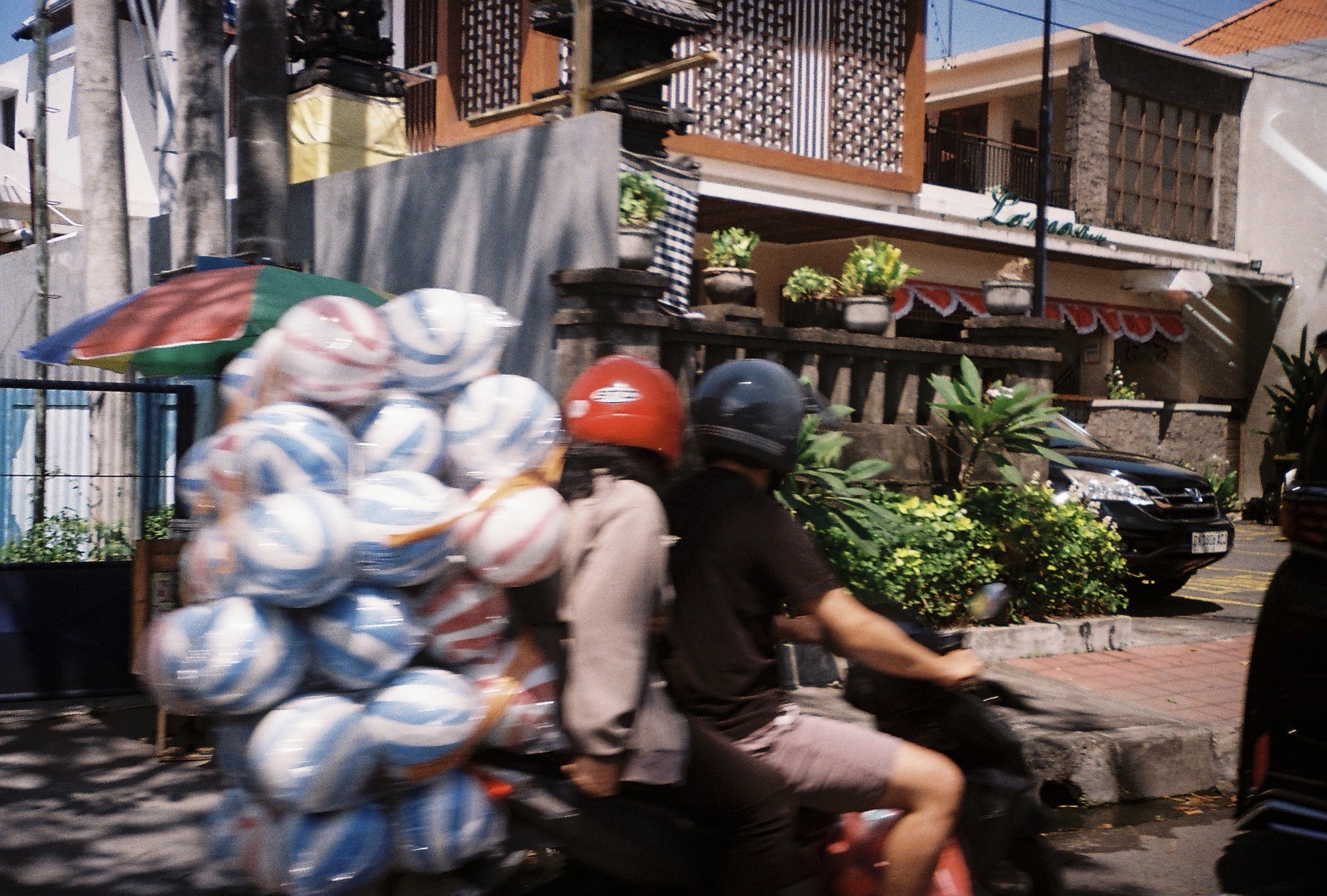 Two helmeted people ride a motion-blurred motorcycle carrying a wide bundle of red, white, and blue striped round objects past roadside plants.