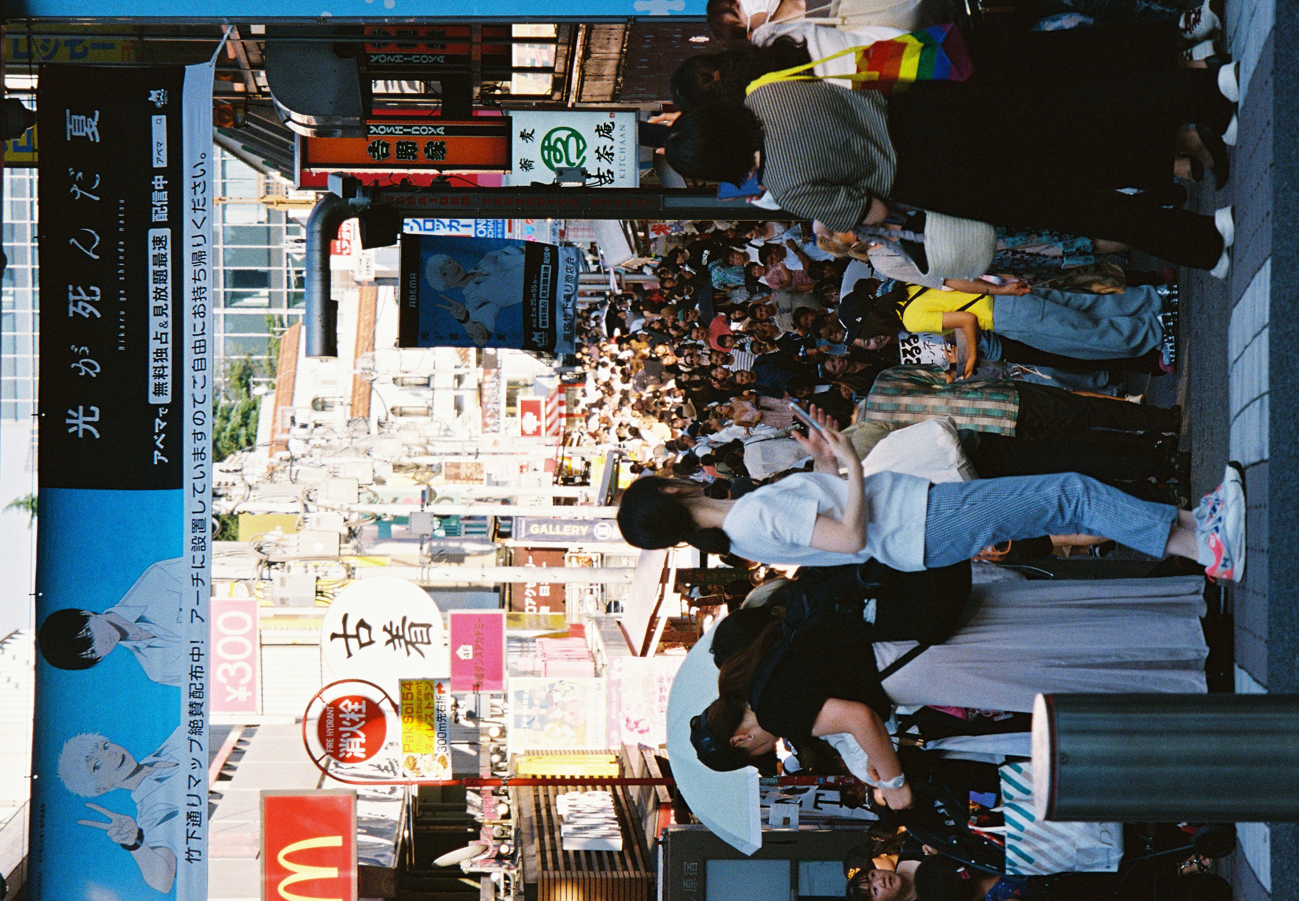 Pedestrians holding a white umbrella and a phone stand near a curb while a dense crowd fills the shopping street behind them.