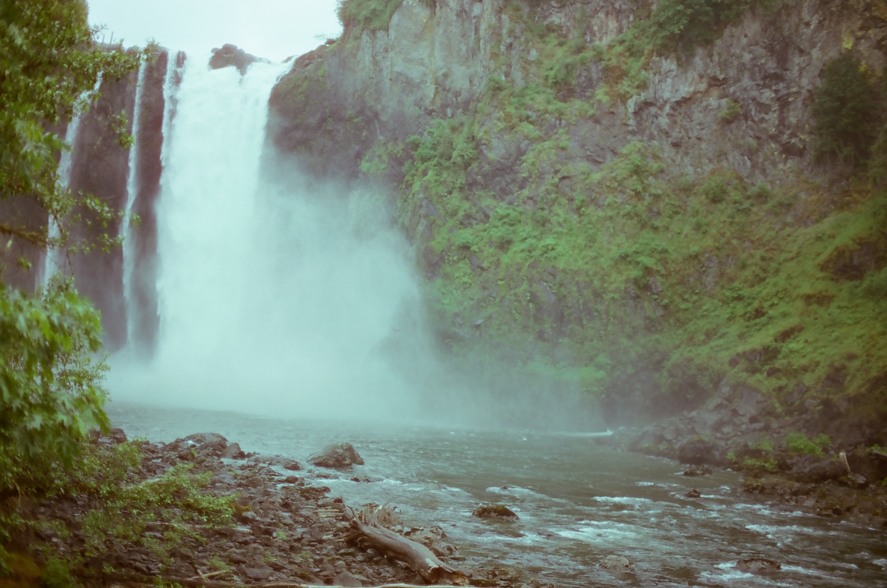 A wide white waterfall plunges between green cliffs into a blue-gray river strewn with stones.