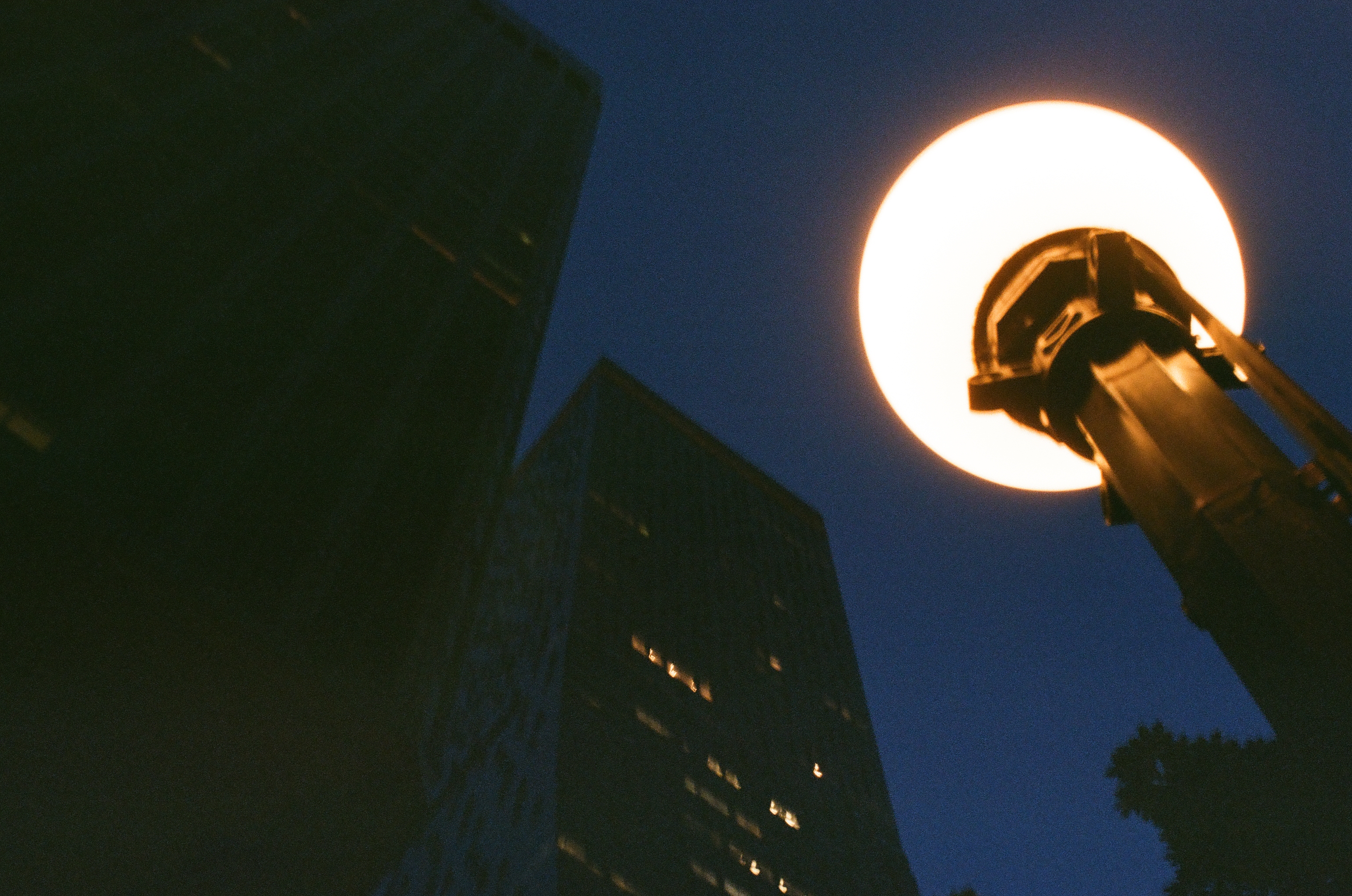 A bright round streetlamp overlaps dark angular towers against a deep blue evening sky.