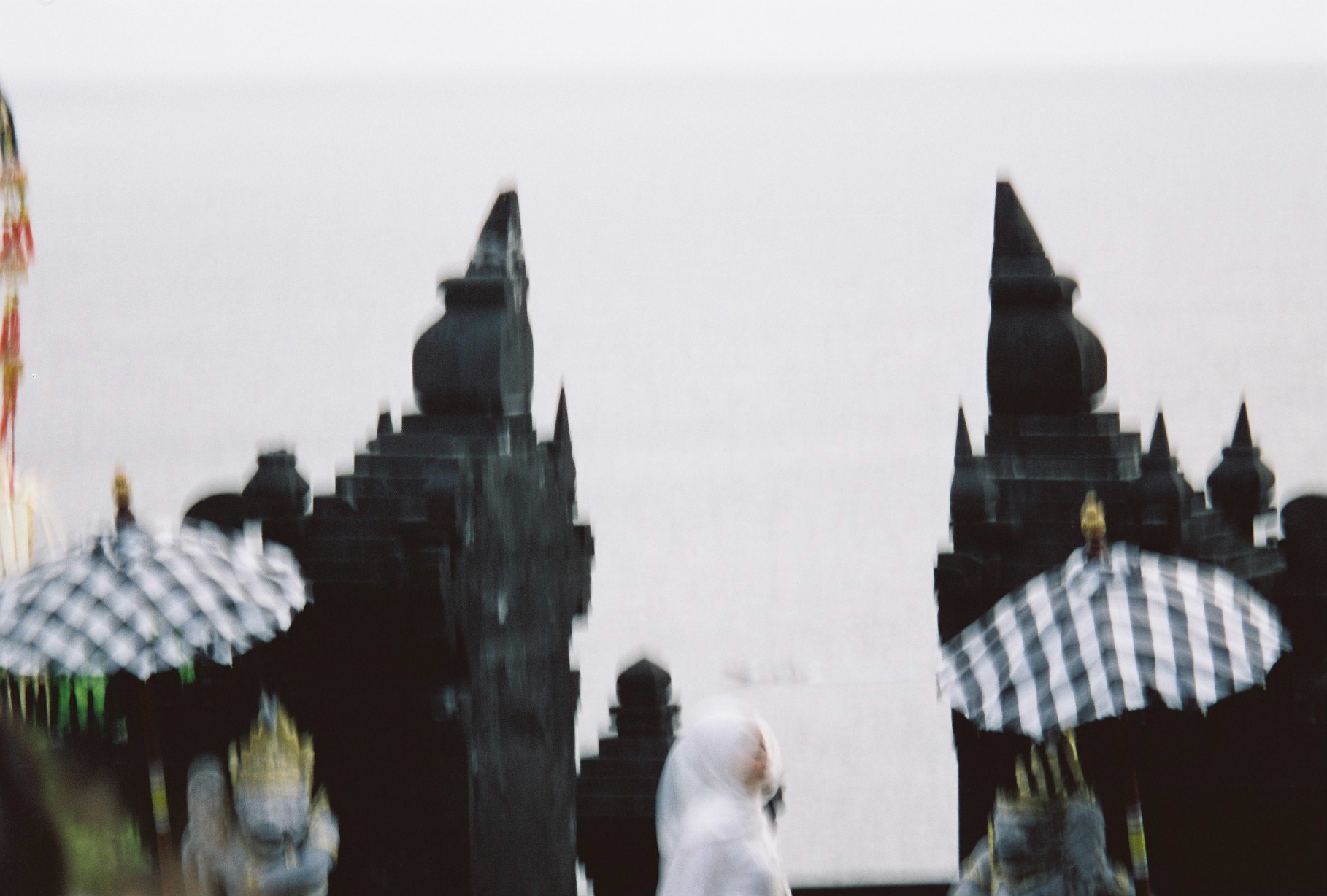 Two dark stepped structures rise against a pale background, with checkered umbrellas and one blurred light-clothed figure below.
