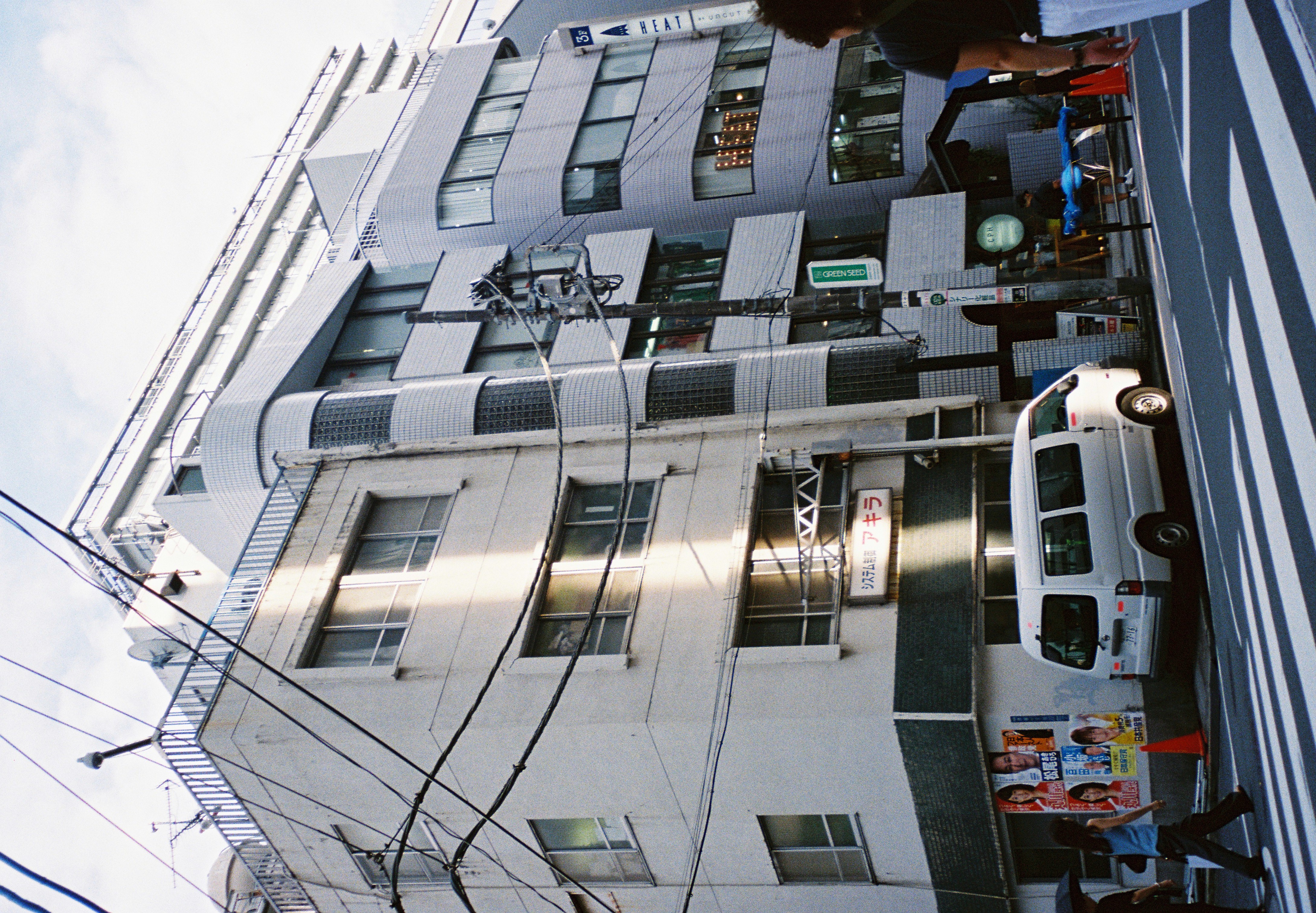 Seen from across the street, the same tiled building and glass-block tower stand beside a crosswalk where pedestrians walk under crossing power lines.