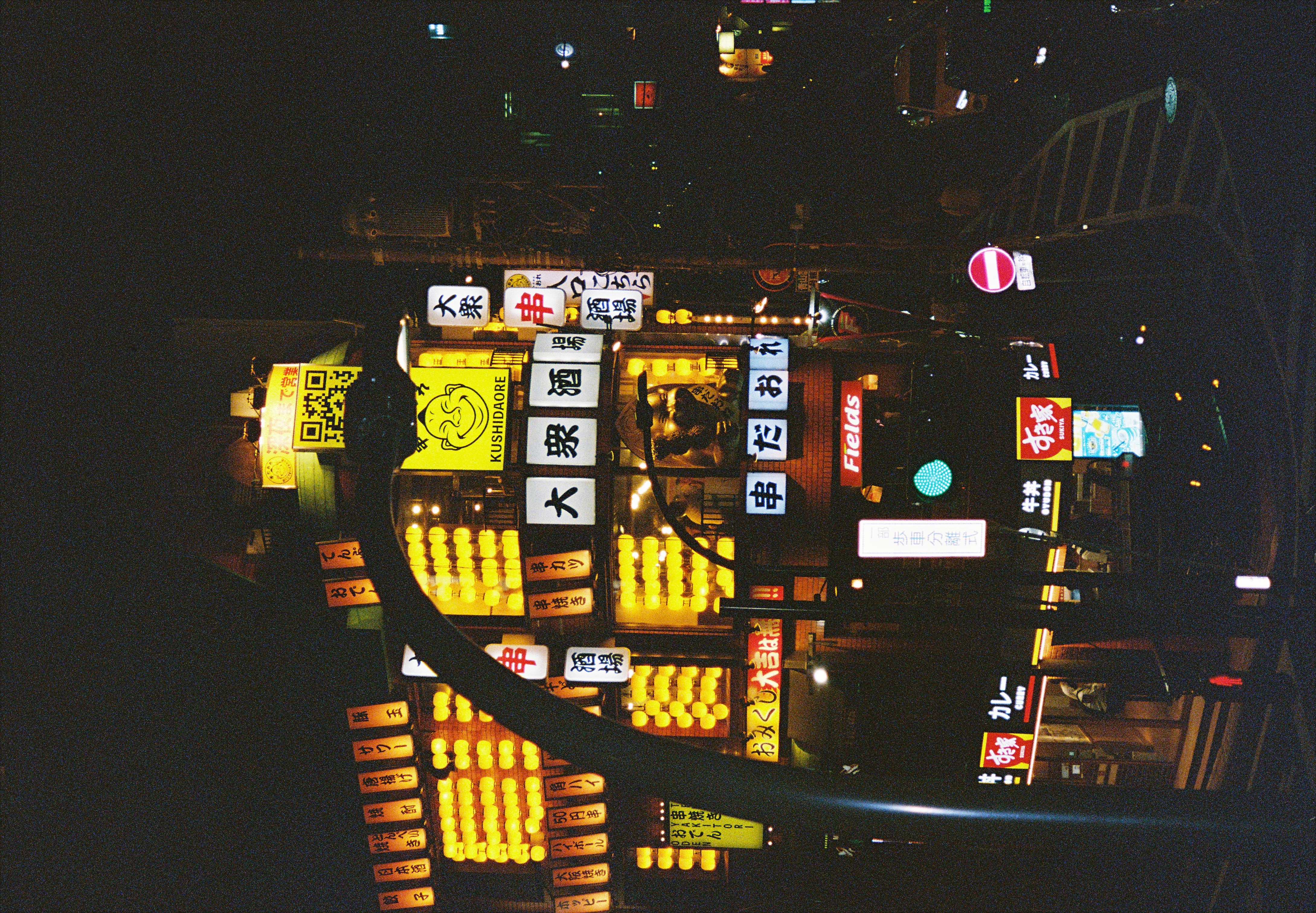 A tall building at night covered in rows of glowing yellow lanterns and stacked signs, with a curved lamppost, green signal, and round red sign in front.