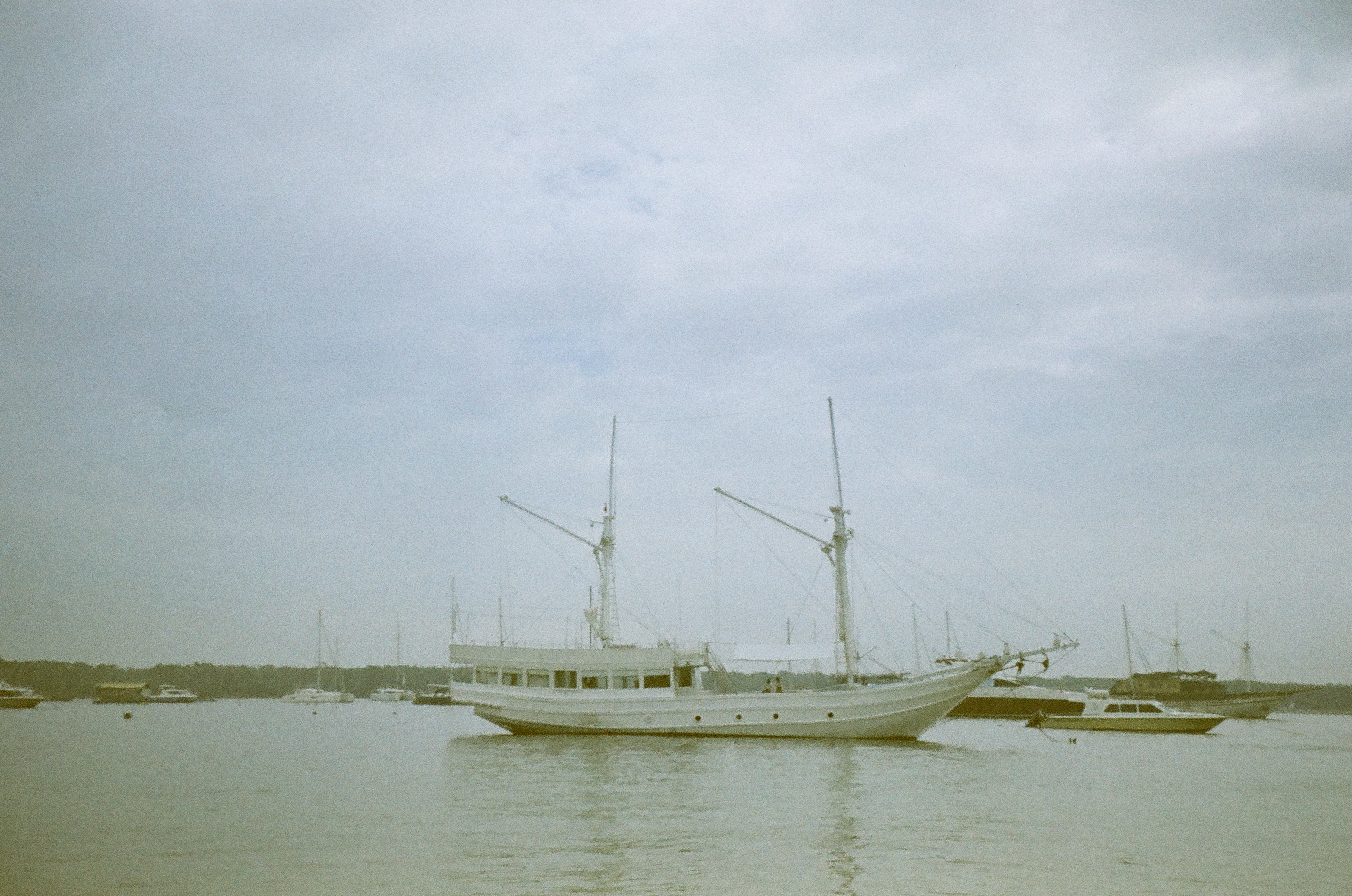 A large two-masted vessel sits among smaller boats on calm water beneath an expansive cloud-filled sky.