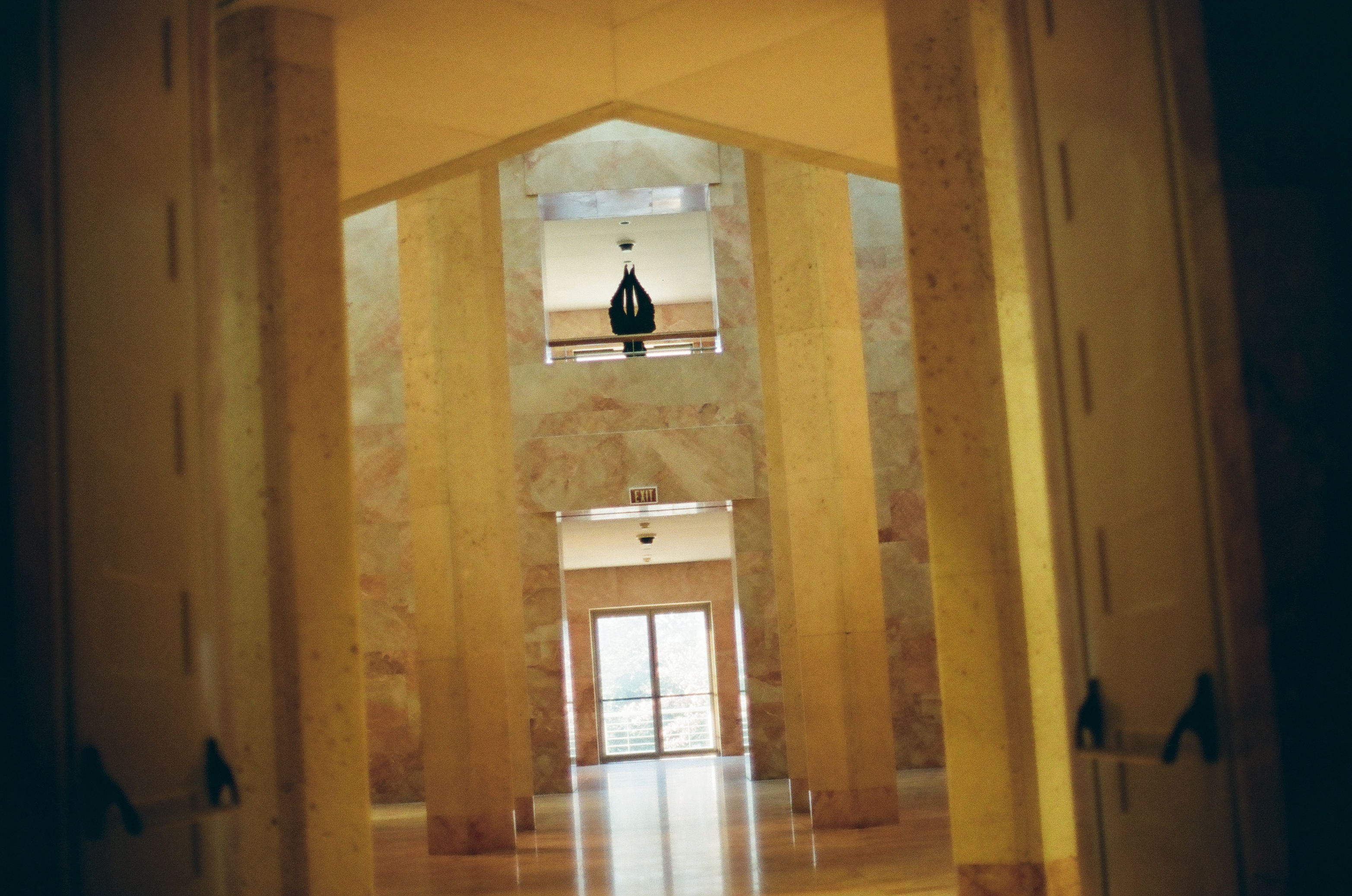 Square stone columns line a warm marble hall toward a glass door, with a dark pointed silhouette hanging in a lit opening above.