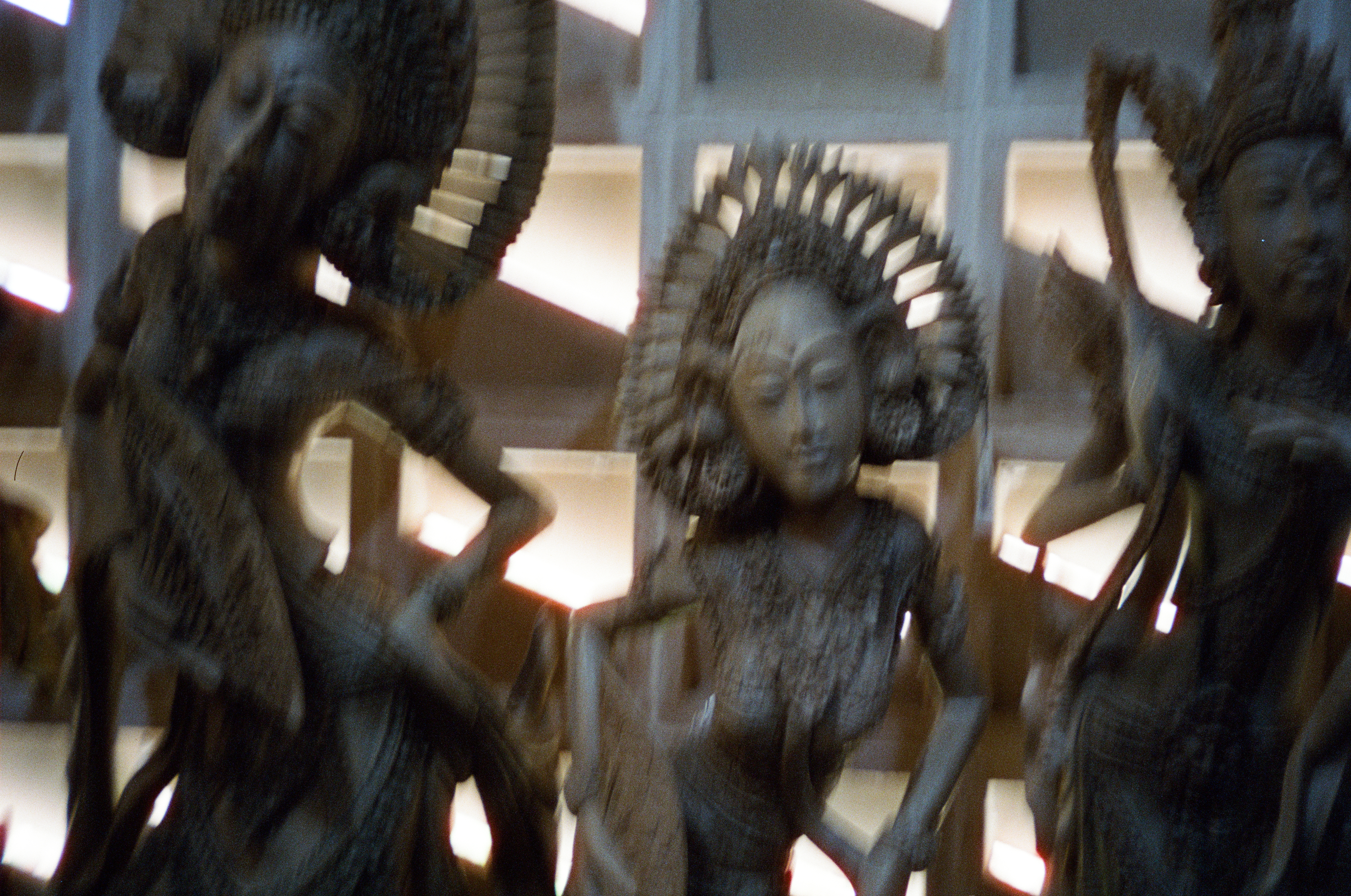 Several dark carved figures with tall headdresses and bent arms stand before bright geometric openings.