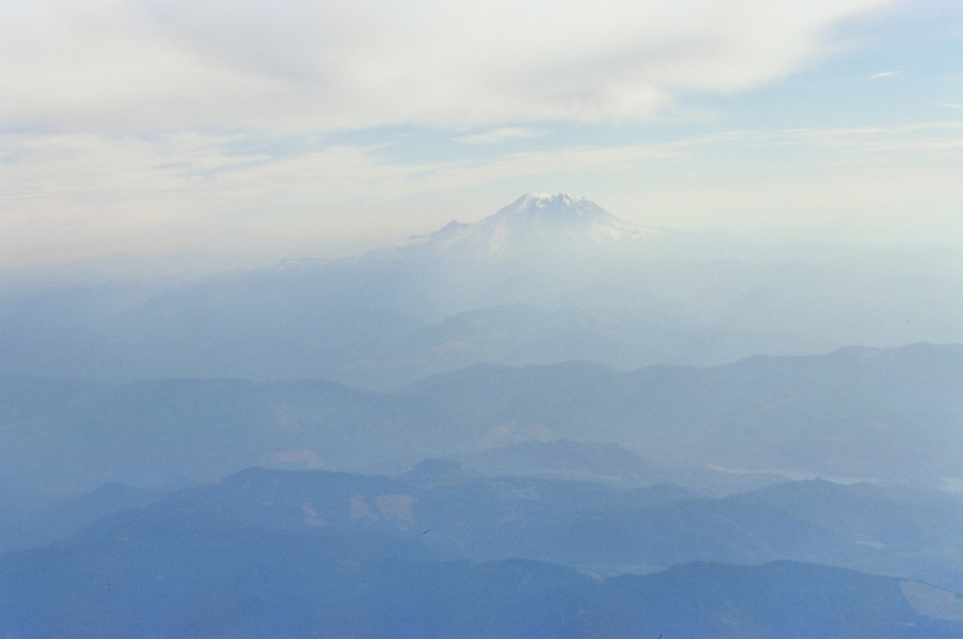 A snow-capped mountain peak stands clear above hazy blue ridgelines, with a faint pale patch resembling water visible in a valley toward the lower right of the frame.