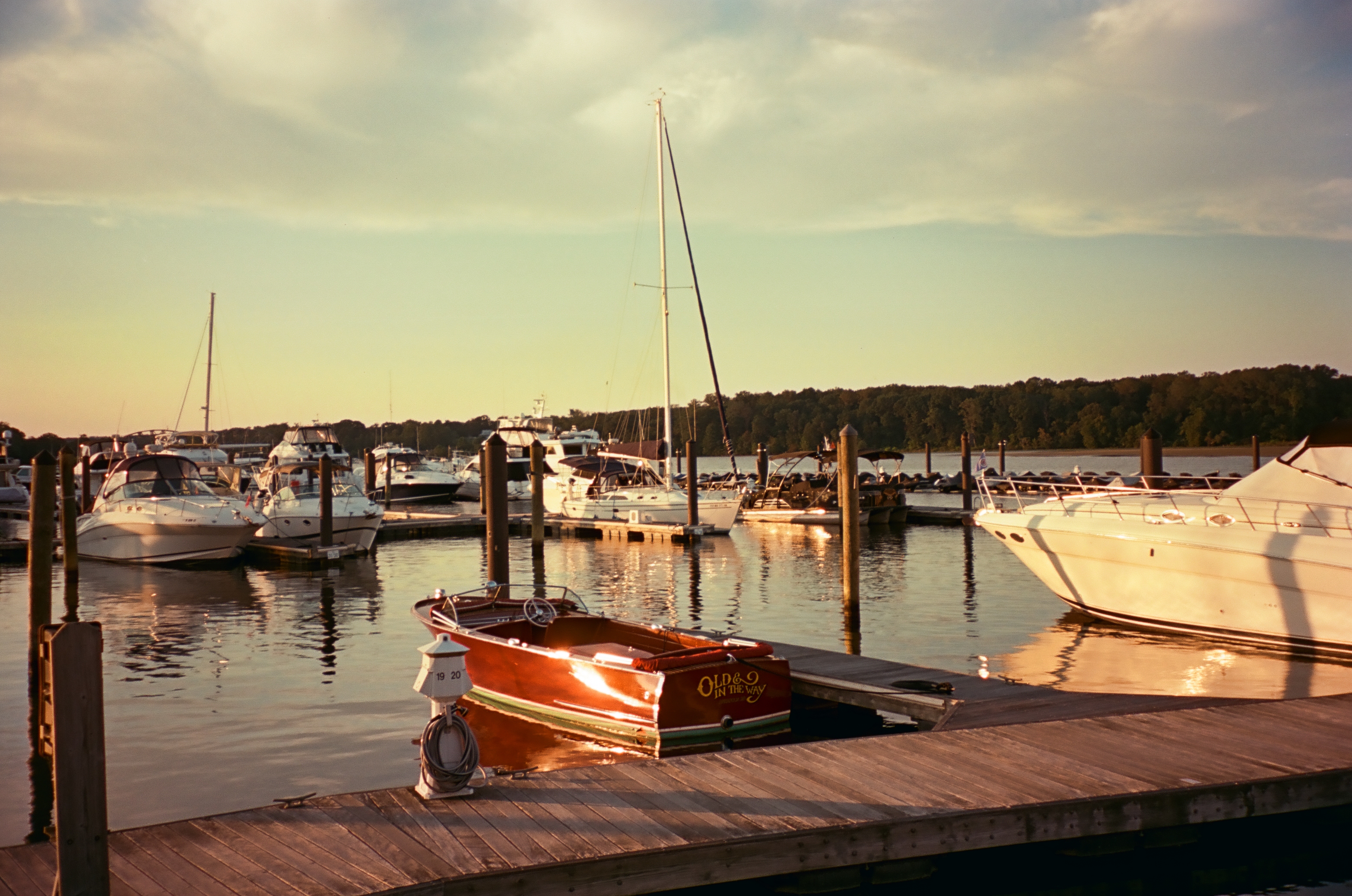A dark red wooden runabout named Old and in the Way is moored at a dock in warm sunset light, with sailboats and cabin cruisers moored beyond.
