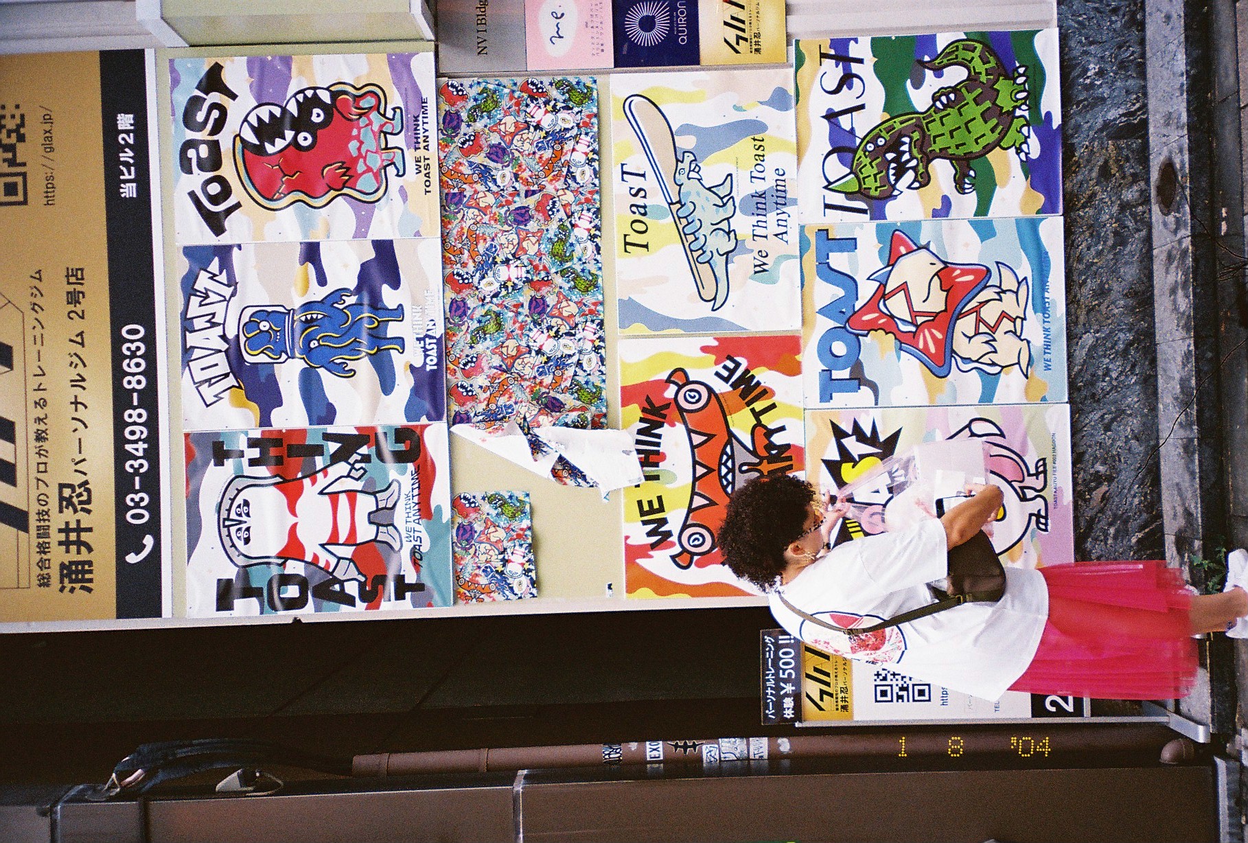 A wall is covered with cartoon character posters repeating the words 'TOAST' and the phrase 'We Think Toast Anytime,' as a person in a pink skirt bends over a bag.