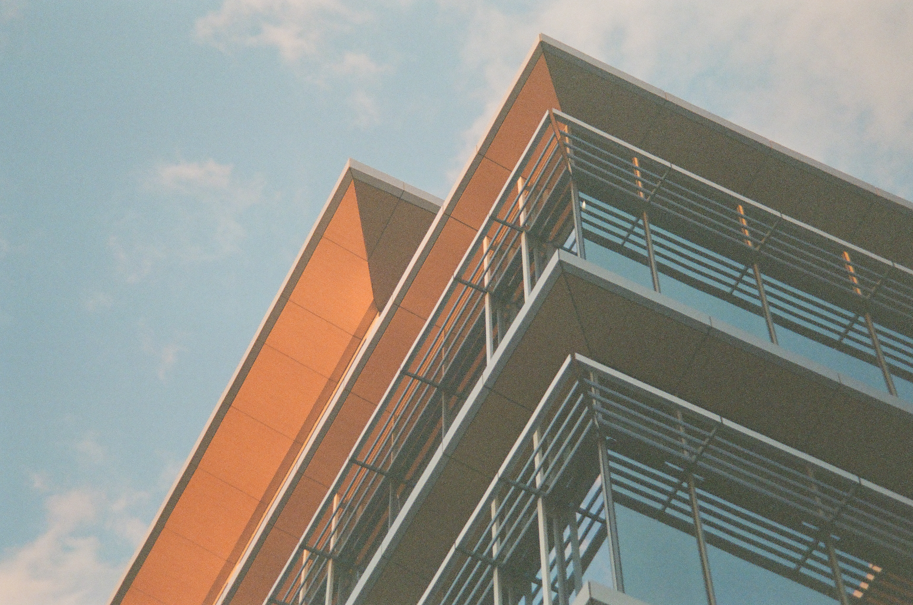A modern glass-and-steel building's corner rises against a blue sky, its overhangs glowing orange beneath stacked gray balcony railings.