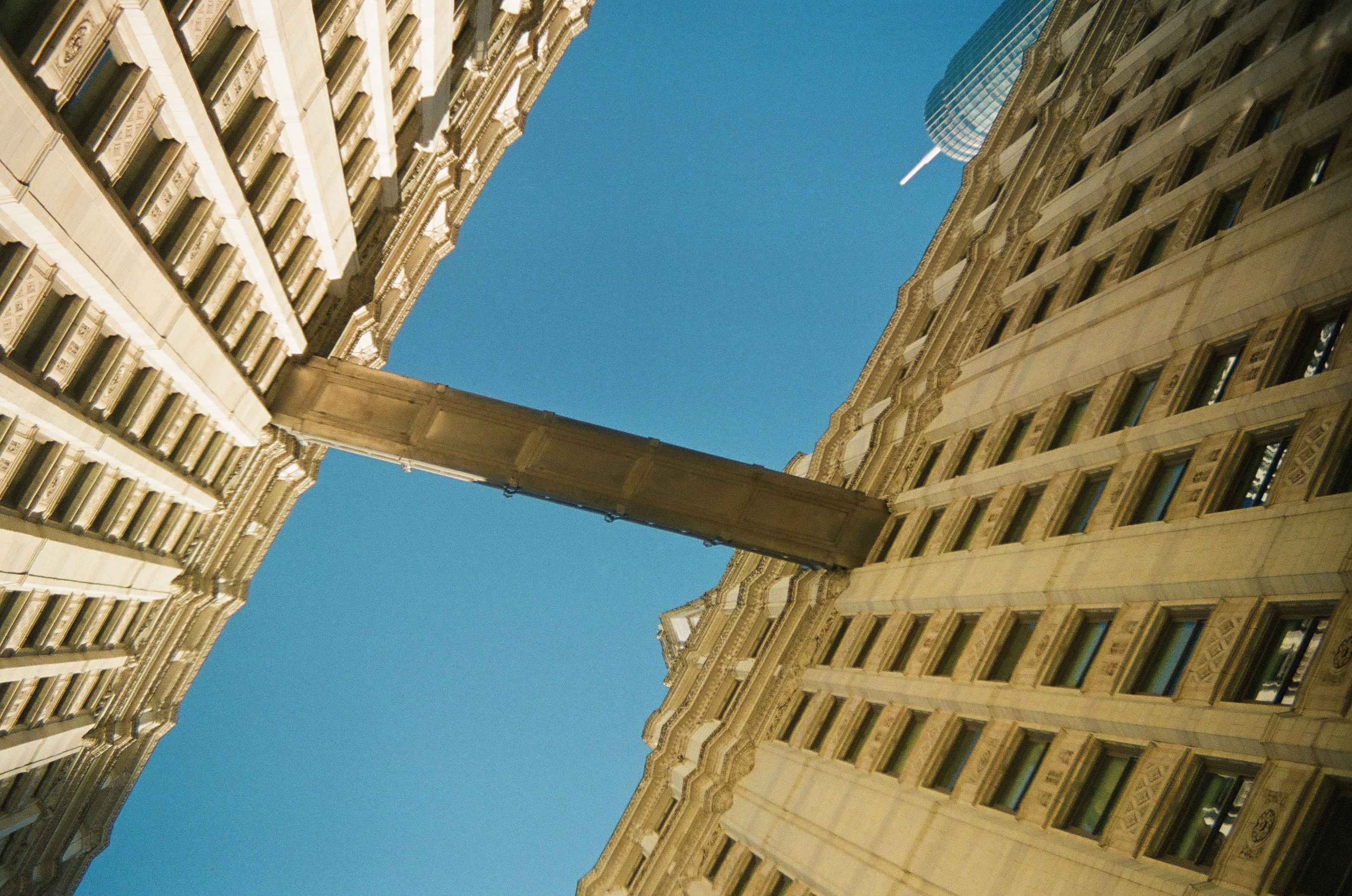 Tall stone facades angle around a narrow overhead bridge beneath a clear blue sky.