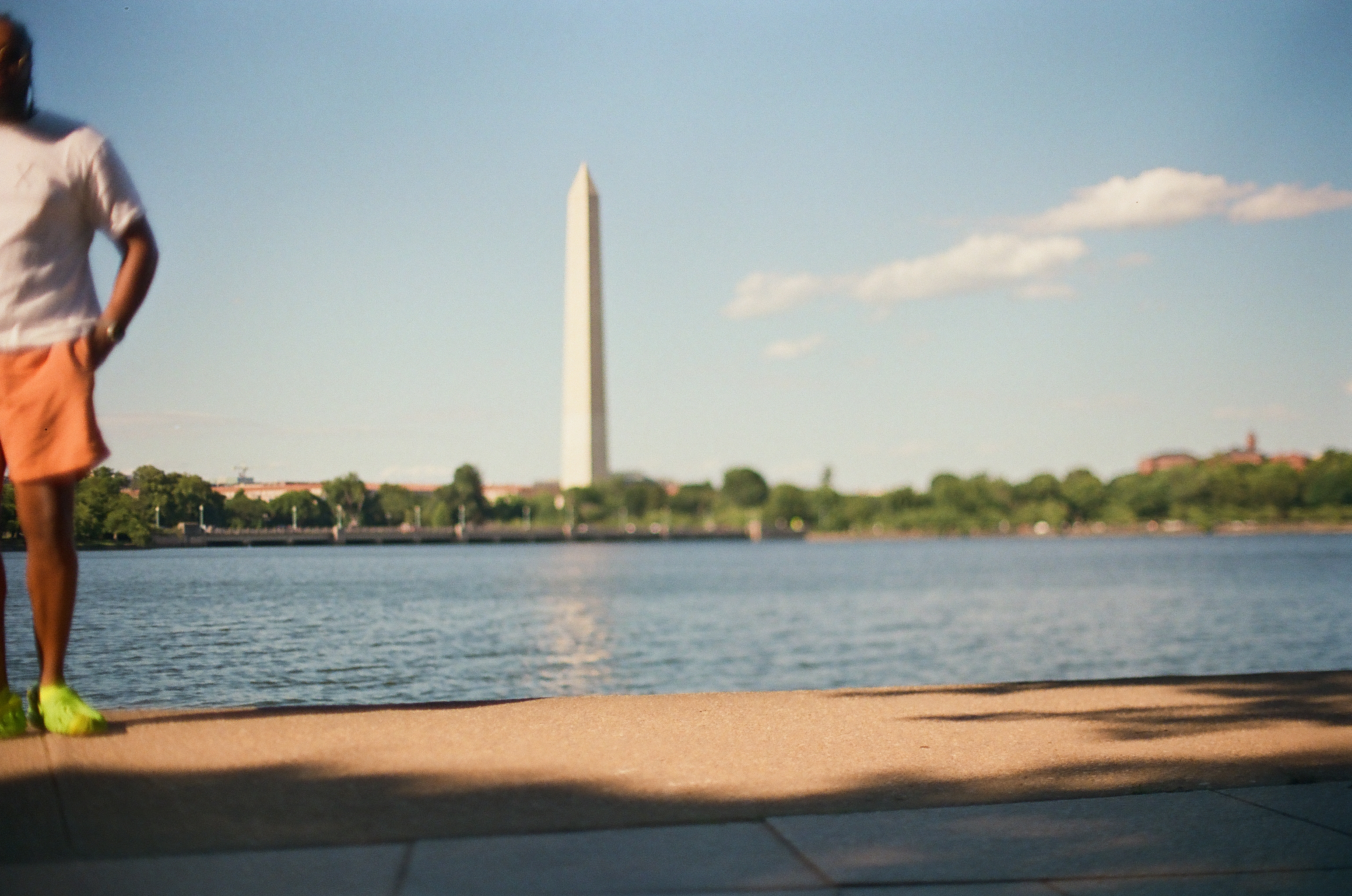 A person stands at the sunlit waterfront edge, hand on hip, facing a tall obelisk across rippling water.