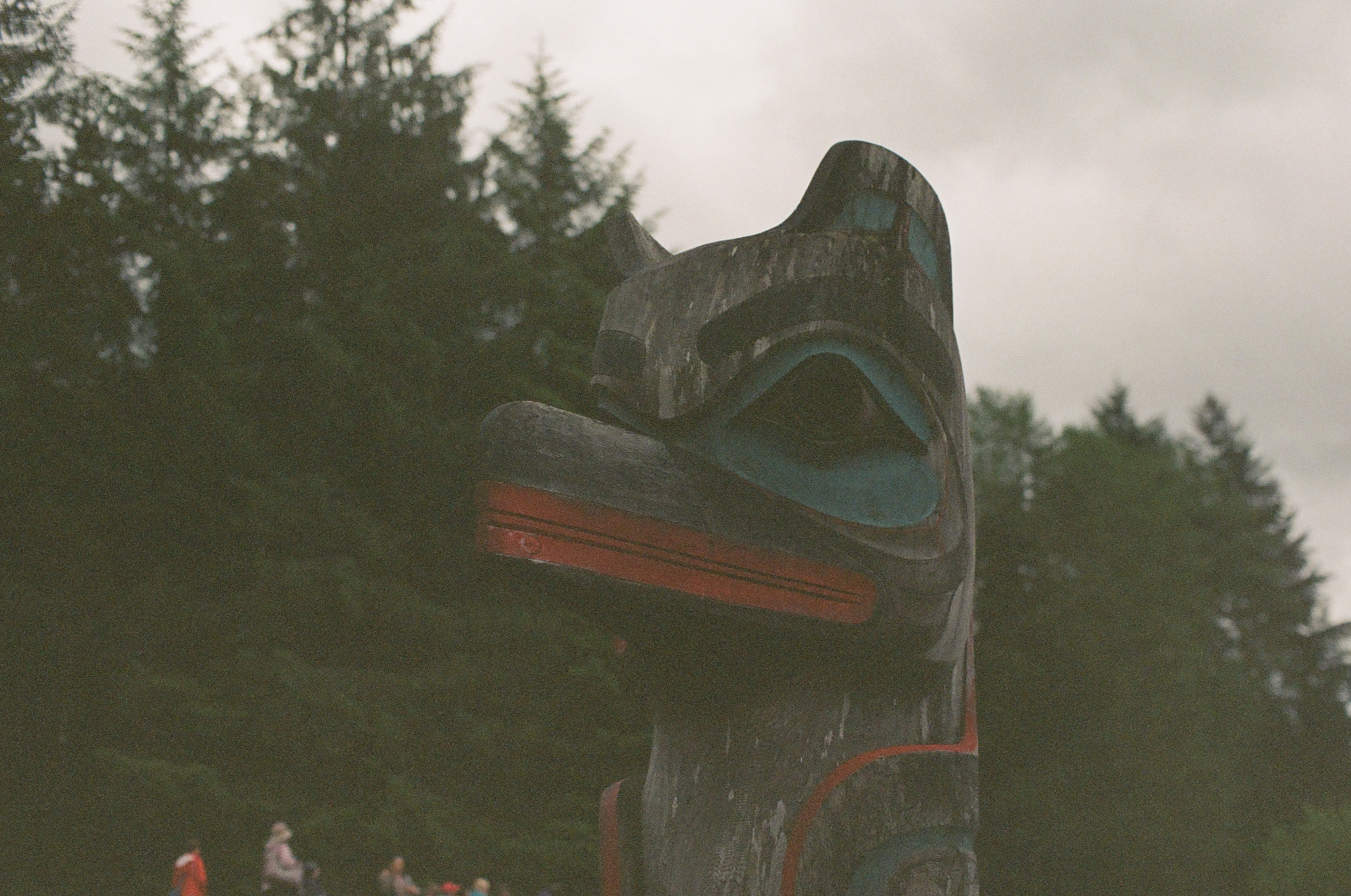 A large dark carved animal-like head rises against evergreen branches above several small figures.