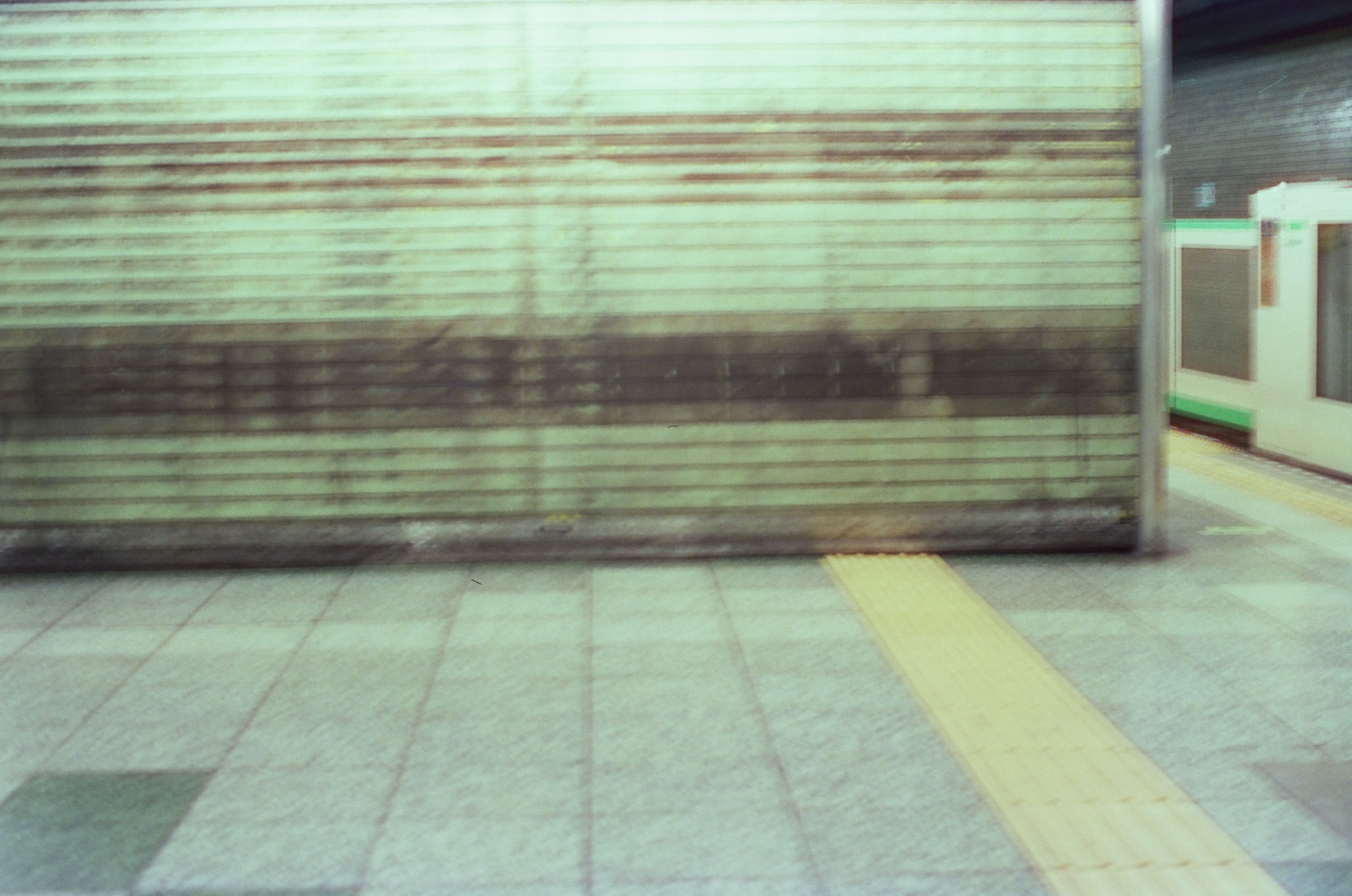 A ribbed metal shutter, tiled pavement, and train edge blur together beneath green fluorescent station light.