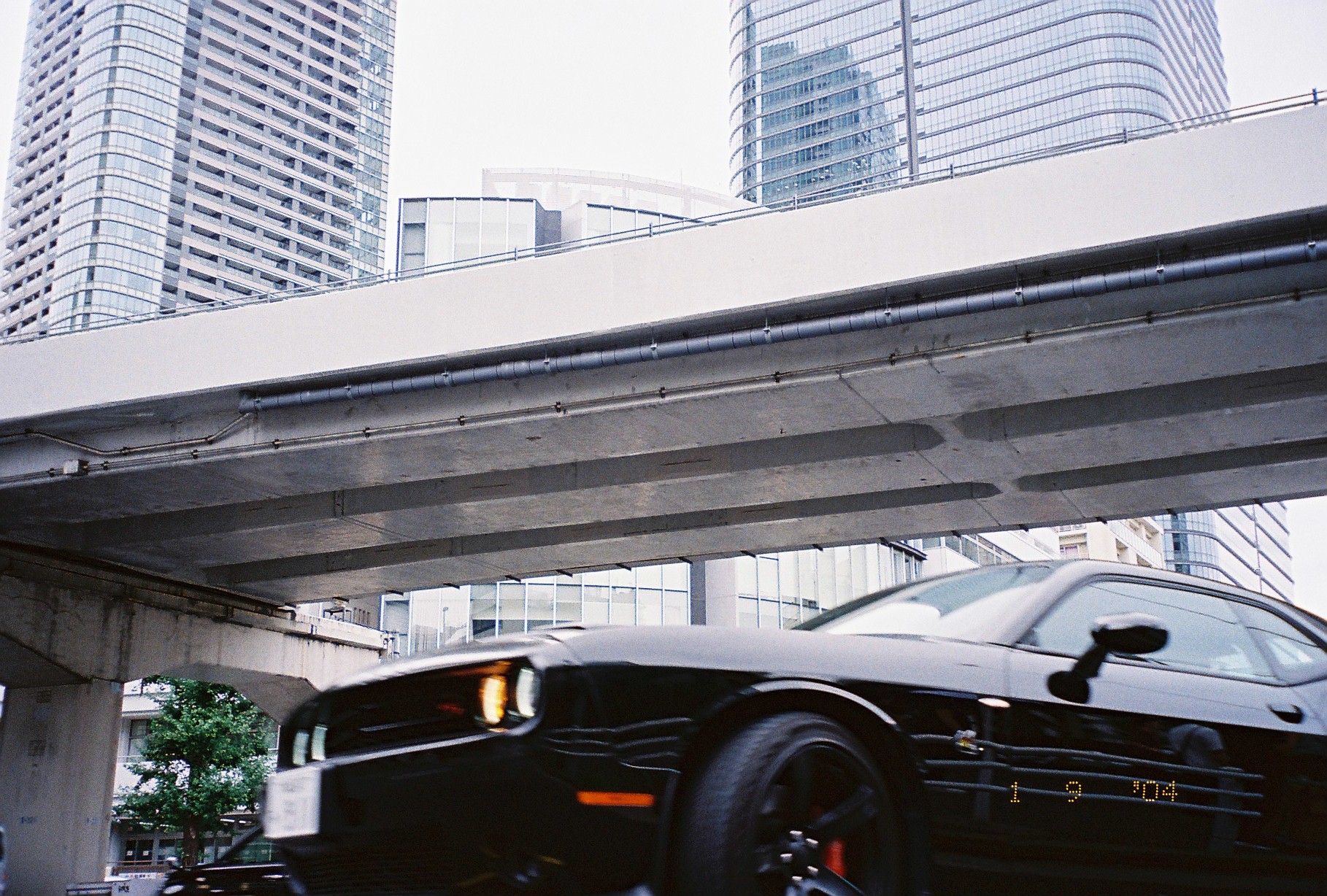 A dark car with a lit headlight passes beneath a concrete expressway overpass, with glass office towers rising behind it.