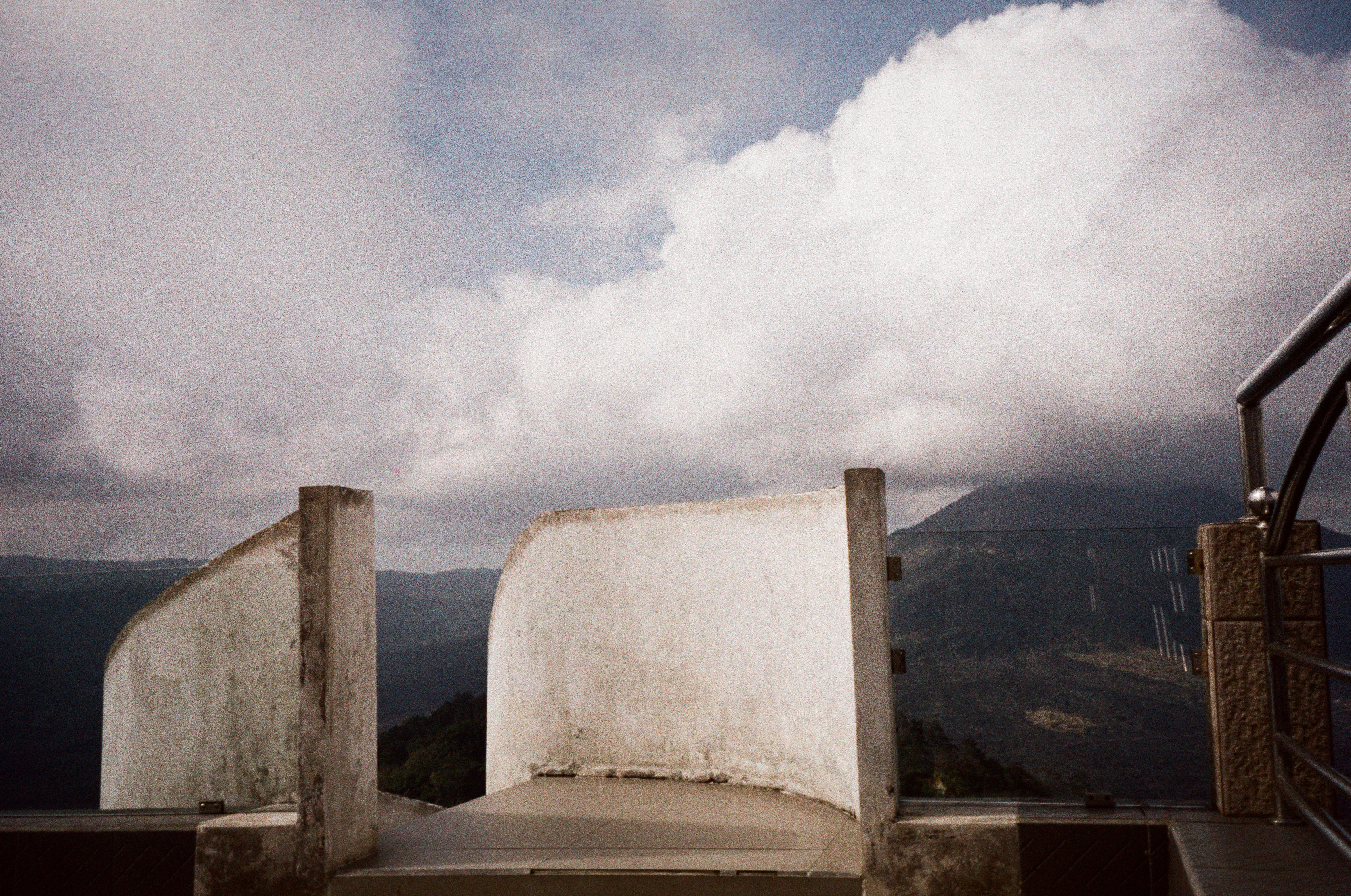 Curved white terrace walls stand below large clouds, with dark ridges behind them and a metal railing at right.